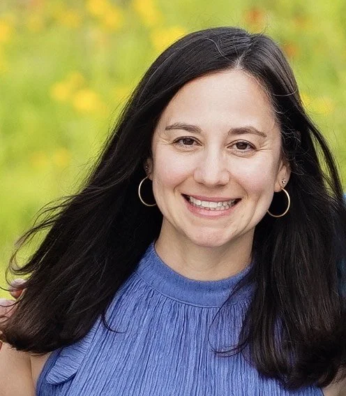 A woman with long dark hair smiling outdoors with a yellow flower background, wearing a blue top and hoop earrings.
