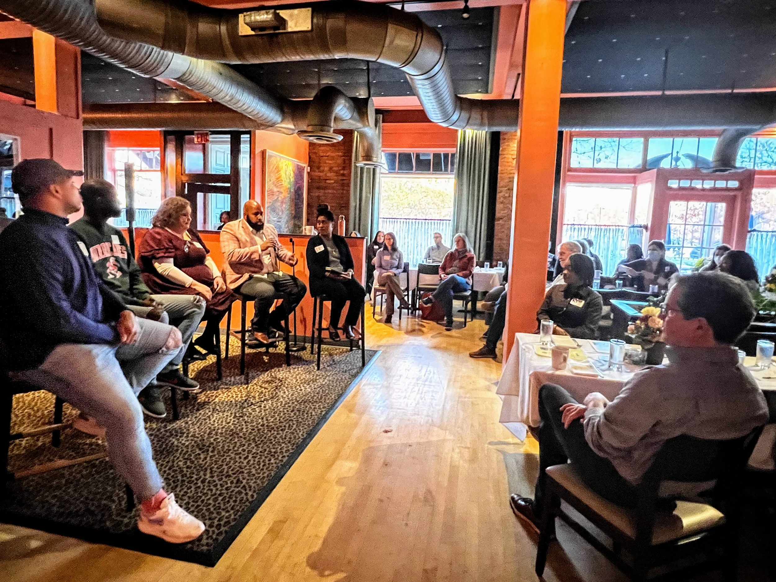 People gathered in a restaurant or event space, sitting in a semi-circle, with a man speaking into a microphone. The setting has warm lighting, large windows letting in natural light, and a rustic, cozy interior with exposed ductwork and wood accents.