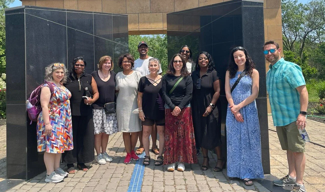 A group of fifteen diverse people smiling for a photo outdoors under a modern archway with trees and greenery in the background.