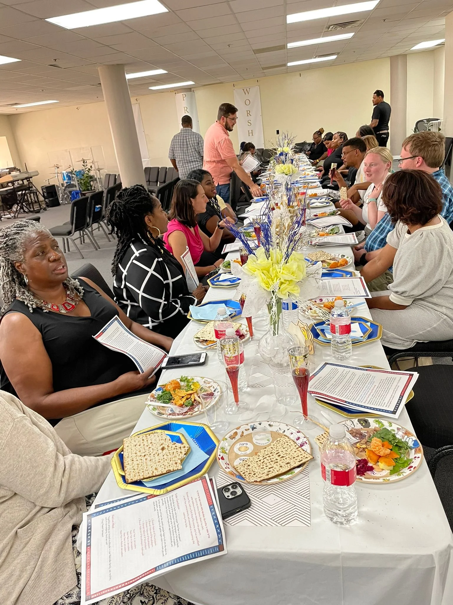 People sitting at a long table during a meal or celebration, with plates of food, drinks, and decorative centerpieces, in a room with banners in the background.