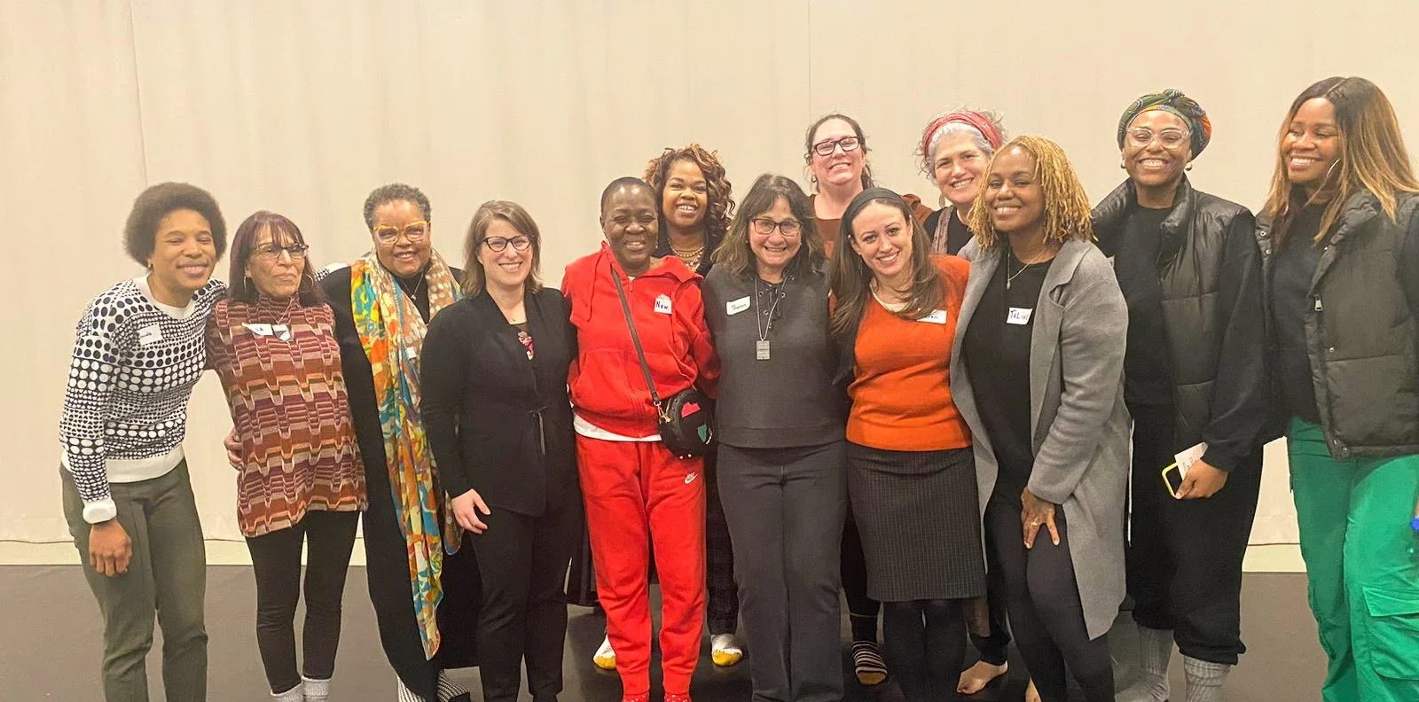 A diverse group of women standing together in a line, smiling for the camera at an indoor event.