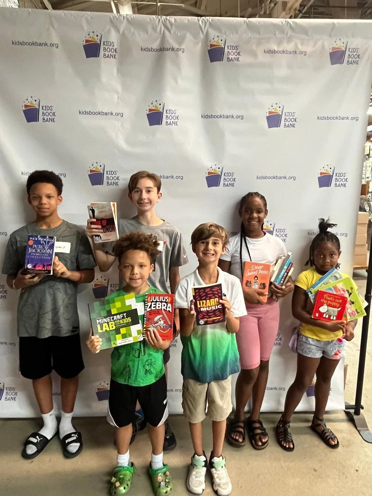 Six children standing in front of a Kids' Book Bank backdrop, holding various books and colorful activity kits, smiling for the photo.