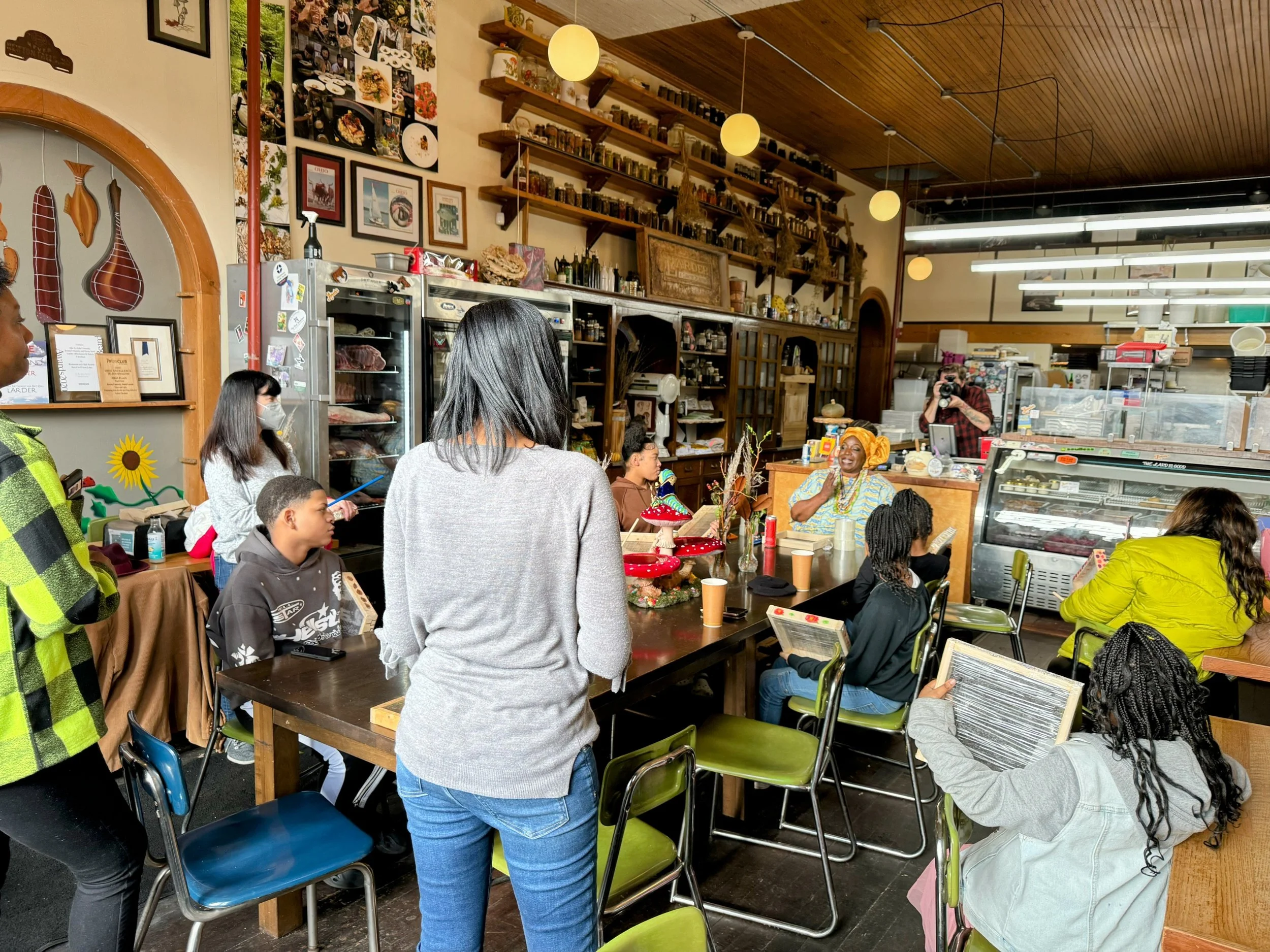 The image shows a lively indoor scene at a restaurant or café with several people. Some individuals are standing, while others are seated, engaging in conversations or using their phones. The interior decor includes wooden shelves filled with jars and pictures on the walls. There are hanging round lights from the wooden ceiling. A woman with a bright yellow headwrap and traditional attire is smiling with her hands clasped. Someone is taking a photo in the background. The atmosphere appears warm and welcoming.