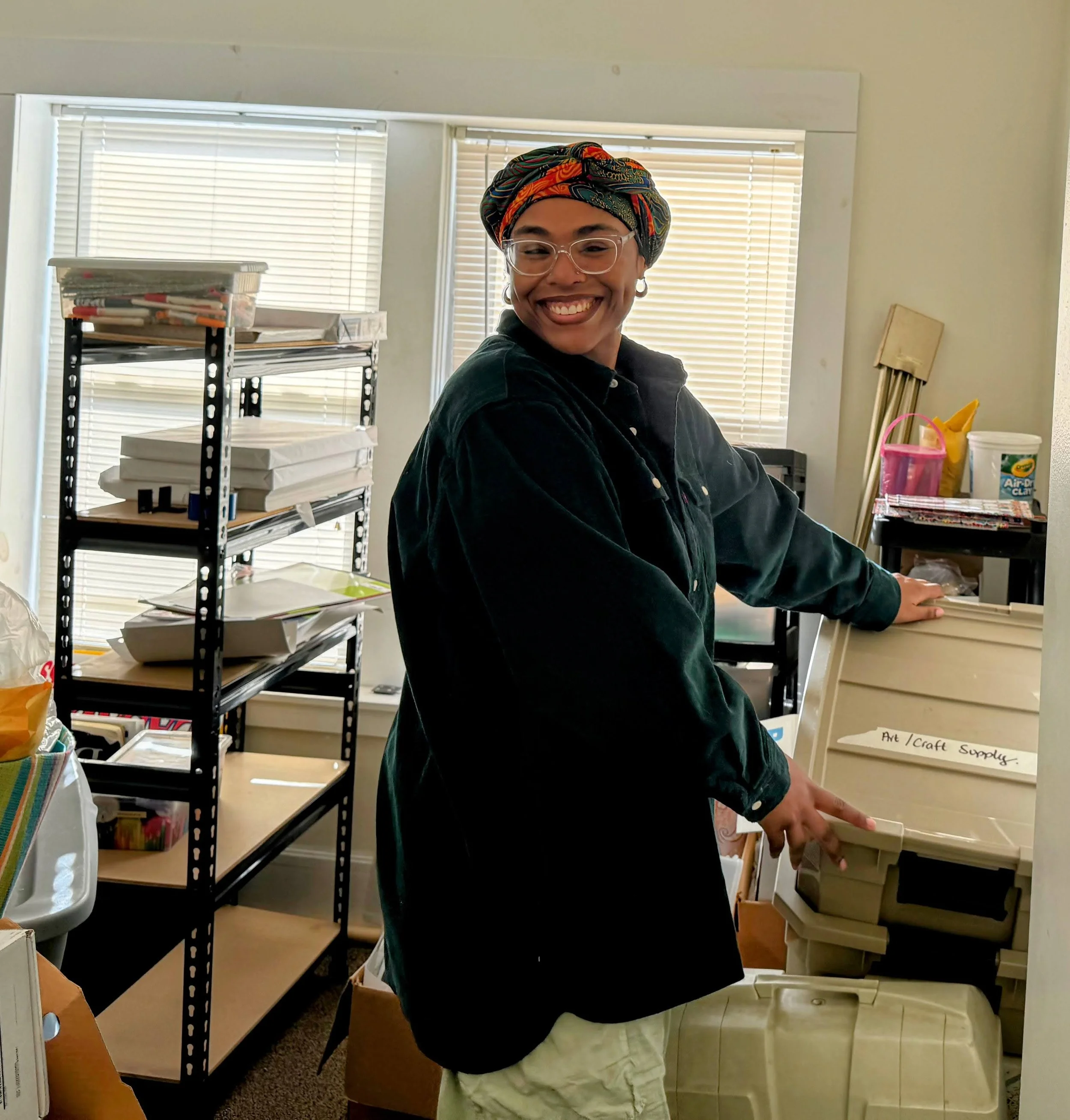 A smiling woman with glasses and a colorful headwrap working in an art and craft supplies area, surrounded by shelves filled with papers, art supplies, and storage bins.