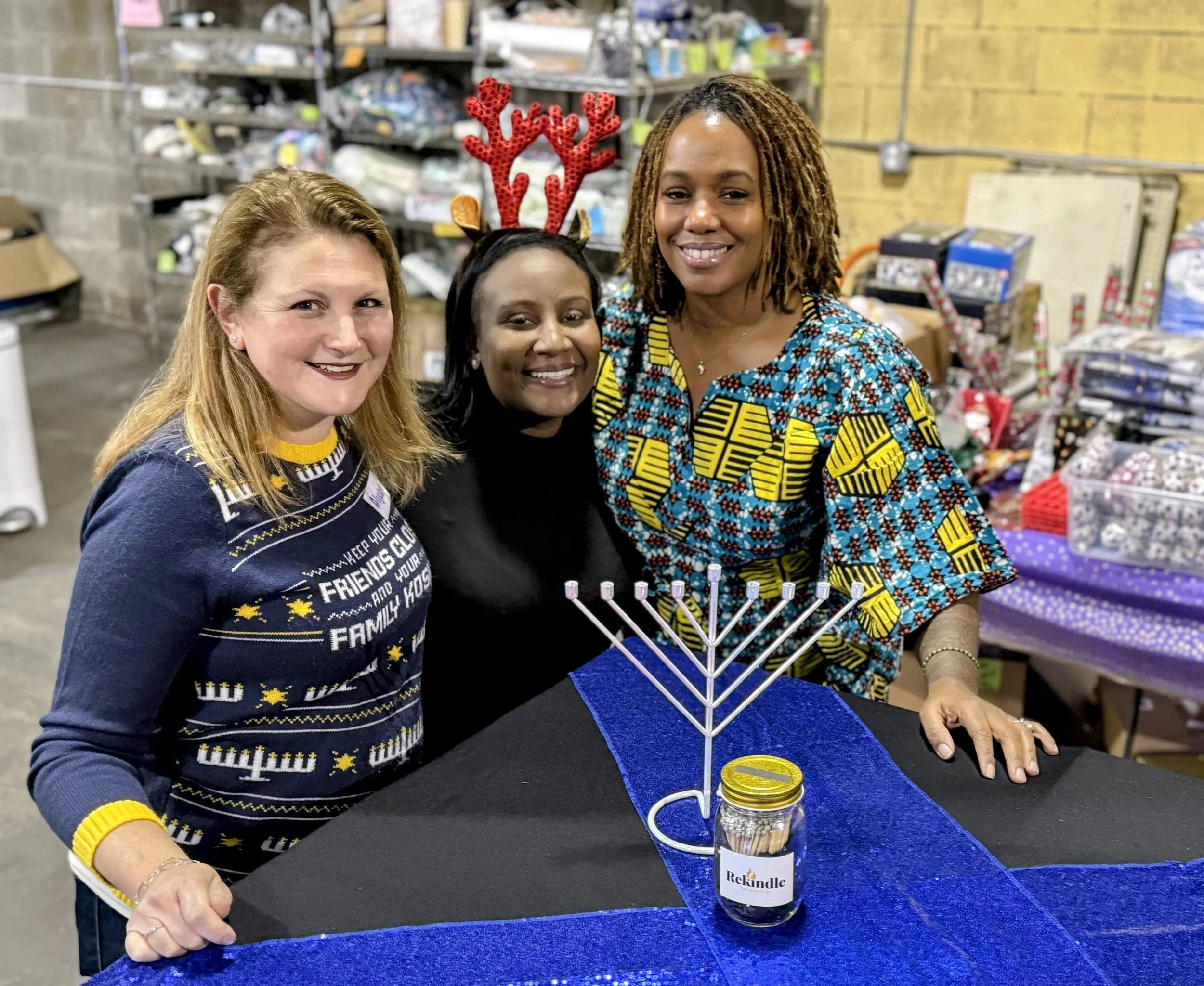Three women smiling at a table with a menorah, some jars, and festive decorations in a warehouse or large room.