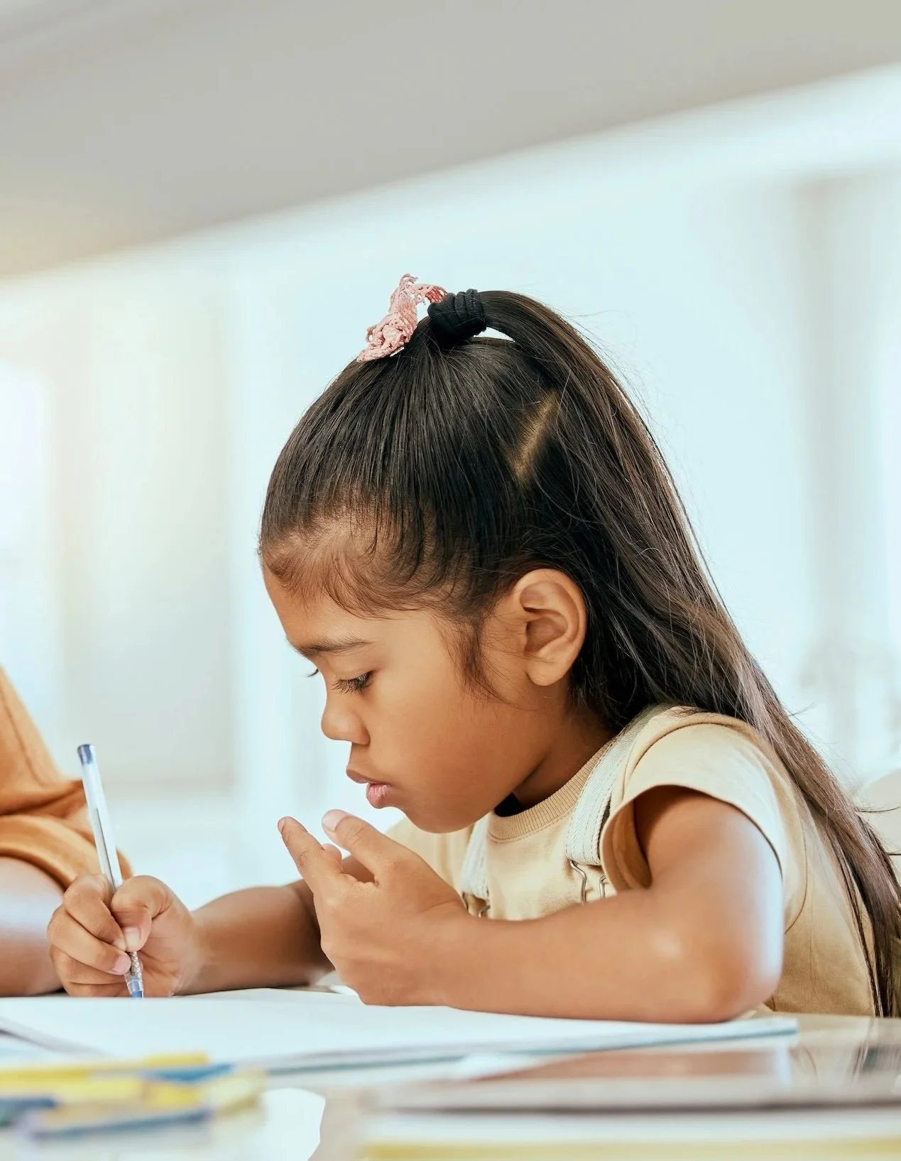 Young girl with long dark hair in a ponytail, wearing a light yellow shirt, focused on writing with a pen on paper at a desk in a bright room.