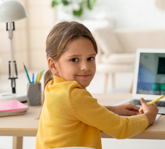 Young girl in a yellow sweater smiling while using a tablet at a desk in a bright room.