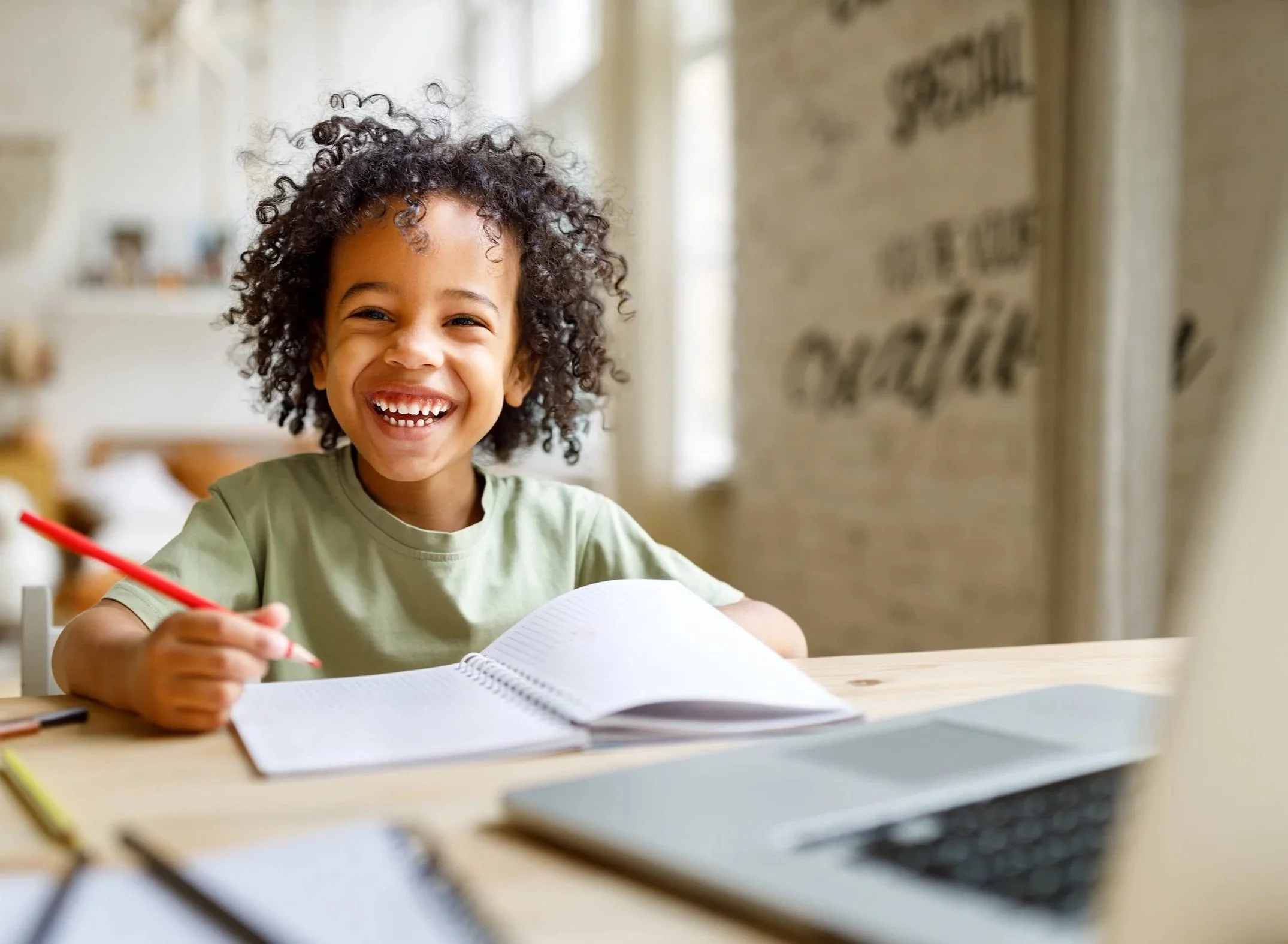 A young boy with curly hair smiling as he writes in a notebook at a desk.