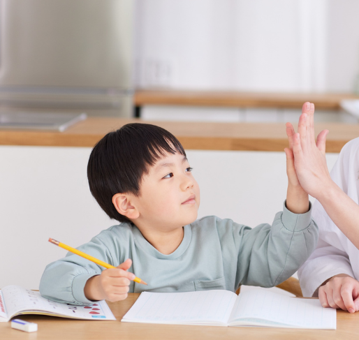 A young boy sitting at a desk with open notebooks, giving a high five to an adult.