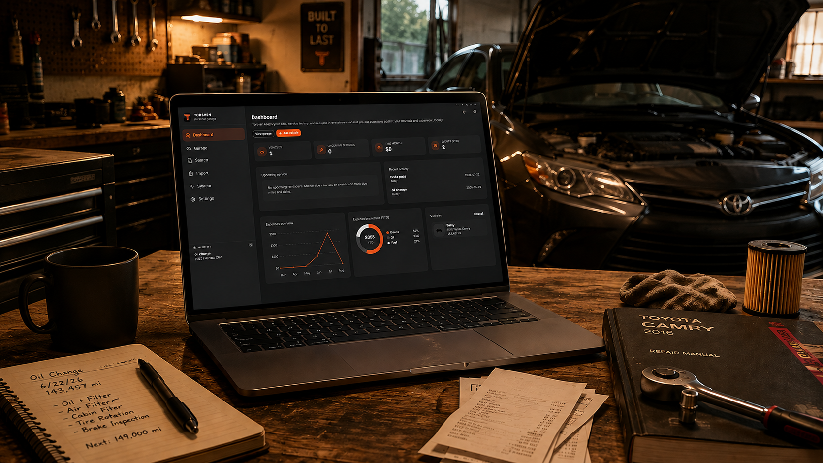 A workspace inside a garage with a laptop showing a car maintenance dashboard, a black coffee mug, a notebook with handwritten notes about oil change, a Toyota Camry repair manual, a tire pressure gauge, and a carbon air filter on a wooden table. A black car with open hood is visible in the background.