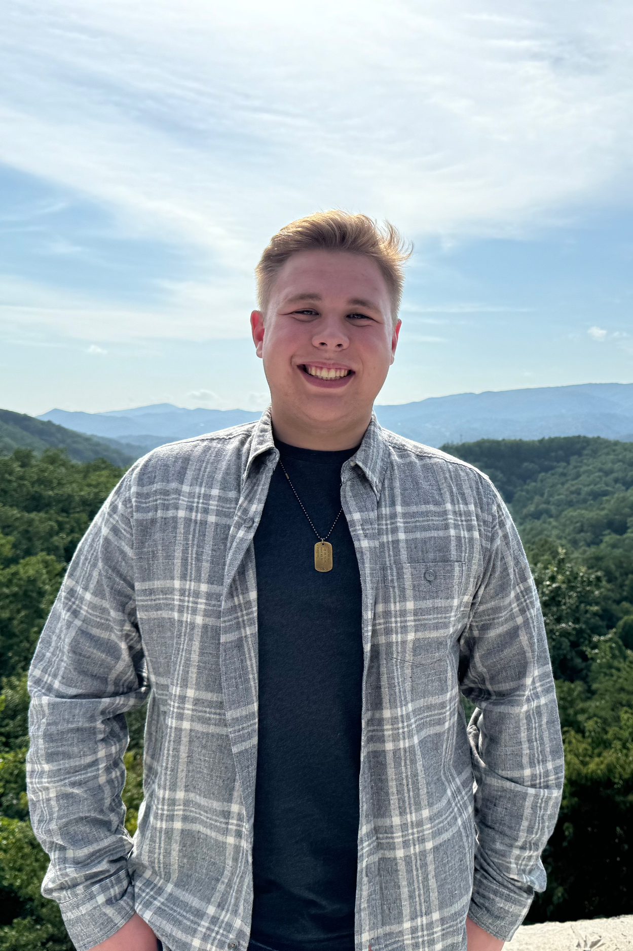 A young man smiling outdoors against a backdrop of green mountains and a partly cloudy sky, wearing a plaid shirt, a black t-shirt, and a dog tag necklace.