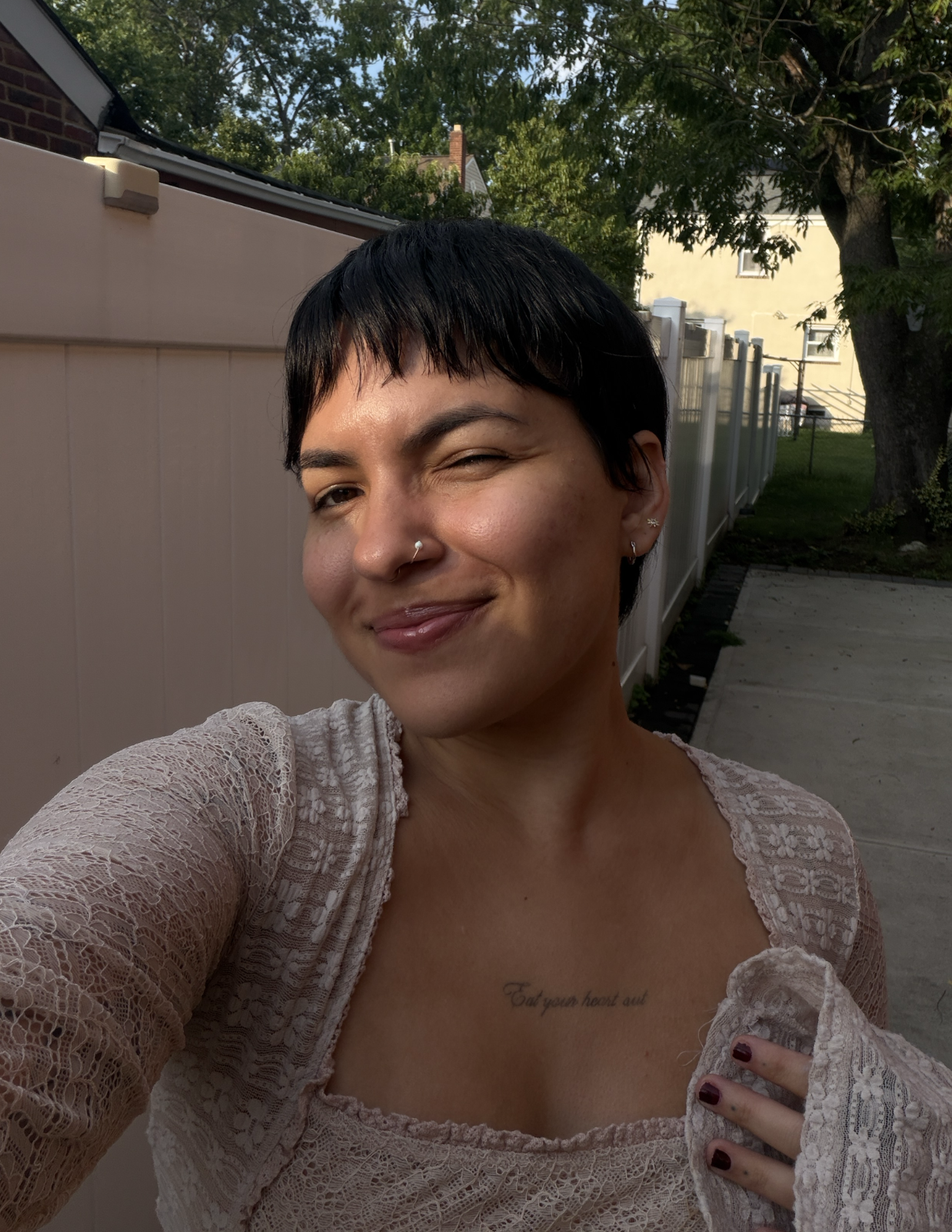 A woman with short dark hair and hoop earrings taking a selfie outside on a sunny day, winking and smiling, wearing a lace top with a tattoo visible on her chest.