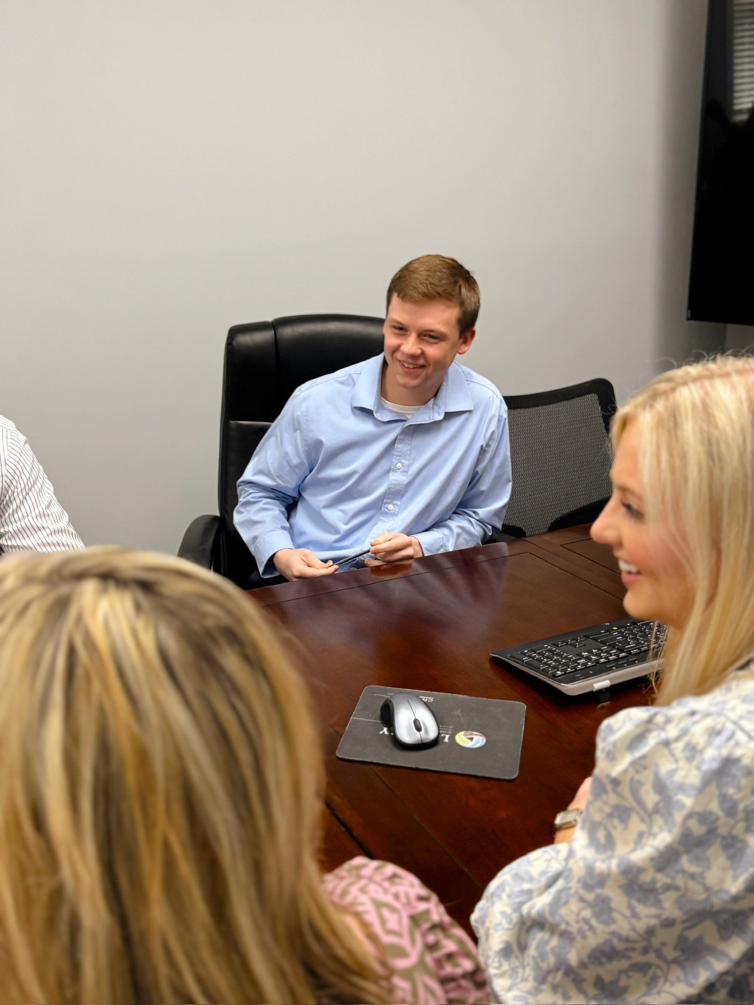 People gathered around a conference table in a meeting room, engaging in a discussion. A young man in a blue shirt is smiling and talking. Laptops, a computer mouse, and a mouse pad are on the table.