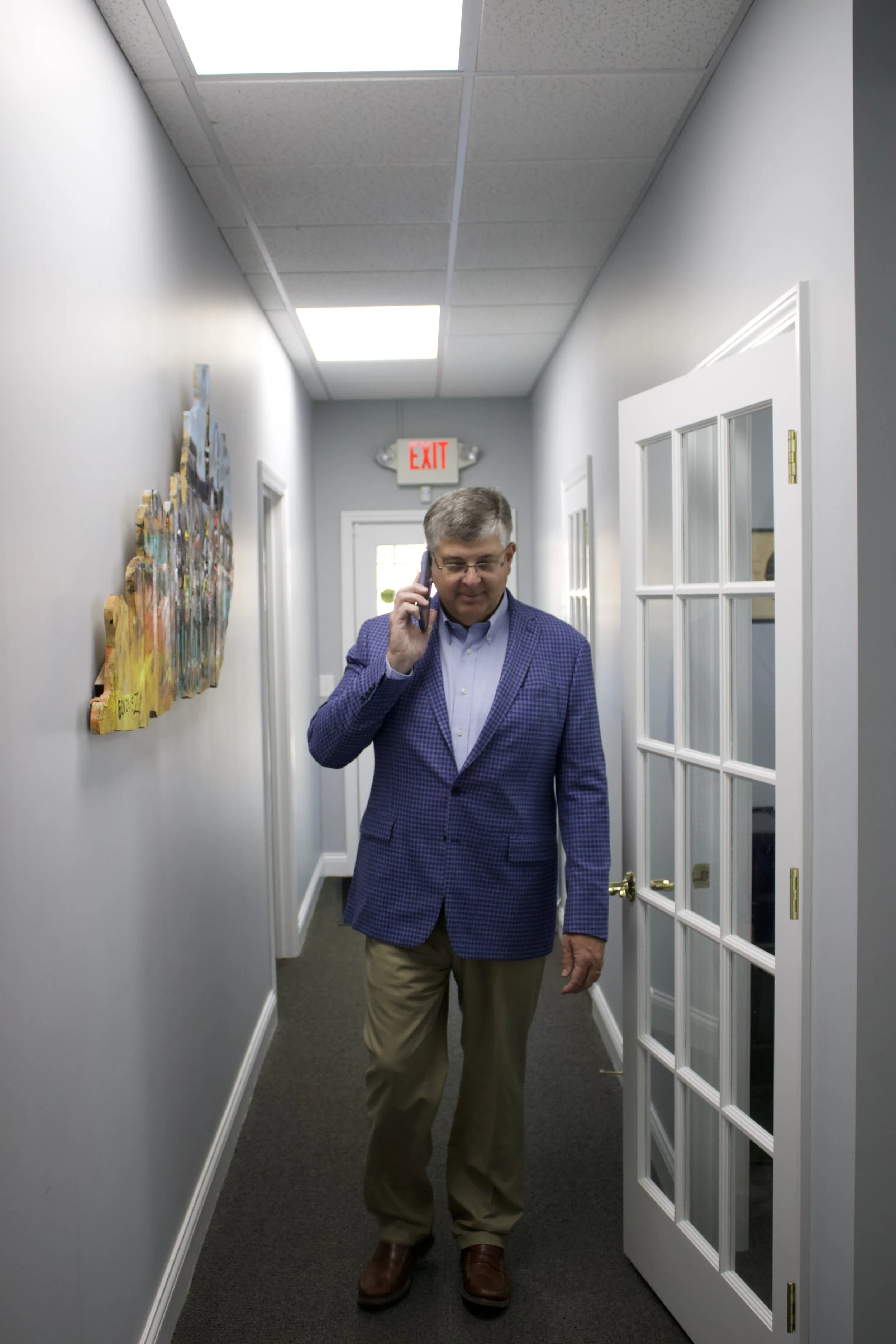 A middle-aged man in a blue plaid blazer, light blue shirt, khaki pants, and brown shoes walking down an office hallway while talking on his cellphone. The hallway has gray walls, white door frames with glass panels on the right, a colorful wall art piece on the left, and the ceiling has fluorescent lights with a visible fire exit sign in the background.