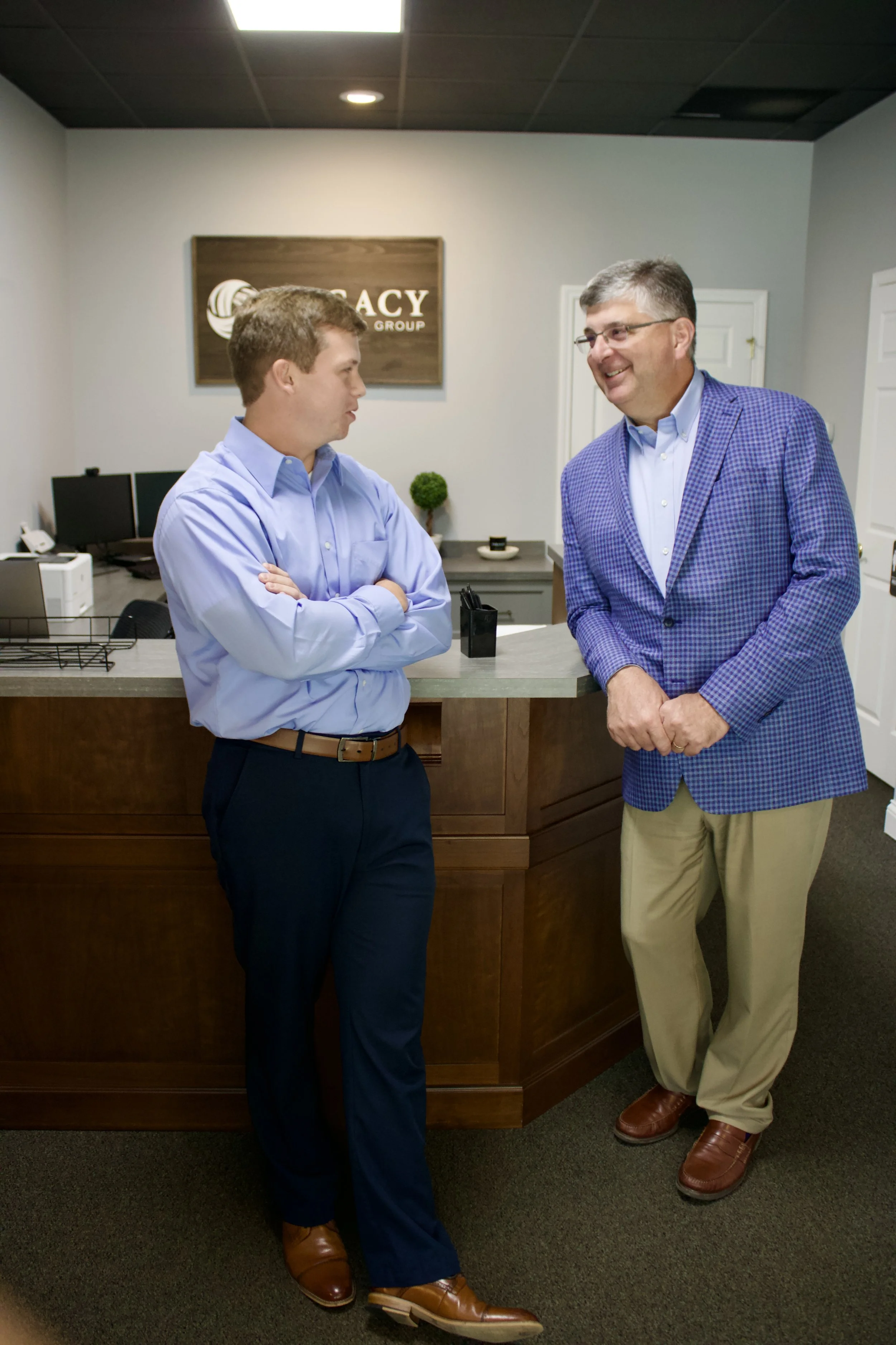 Two men are engaged in conversation in an office reception area. The man on the left is wearing a light blue shirt, dark pants, and brown shoes. The man on the right is wearing a blue checkered blazer, light beige pants, and brown shoes. There is a reception desk with computer monitors and a sign on the wall behind them that reads ‘EMERGENCY GROUP’. The room has white walls, a dark ceiling, and a small decorative plant on the reception desk.