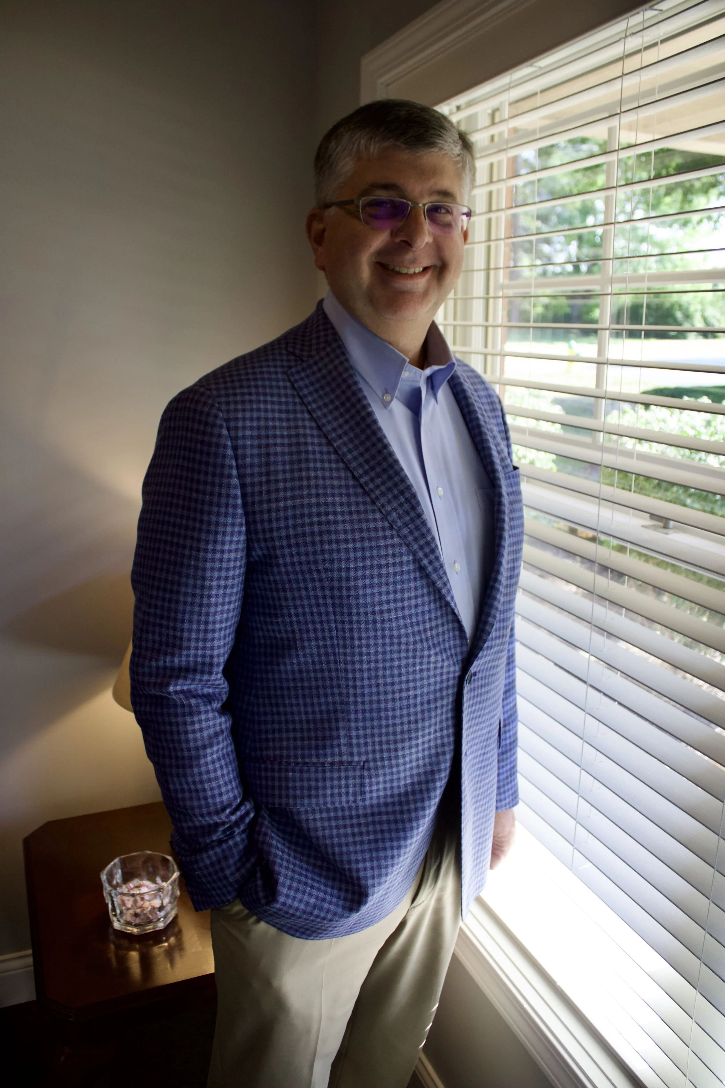 A smiling man in glasses with purple tint, wearing a blue checkered blazer and light-colored pants, standing by a window with blinds in a brightly lit room.