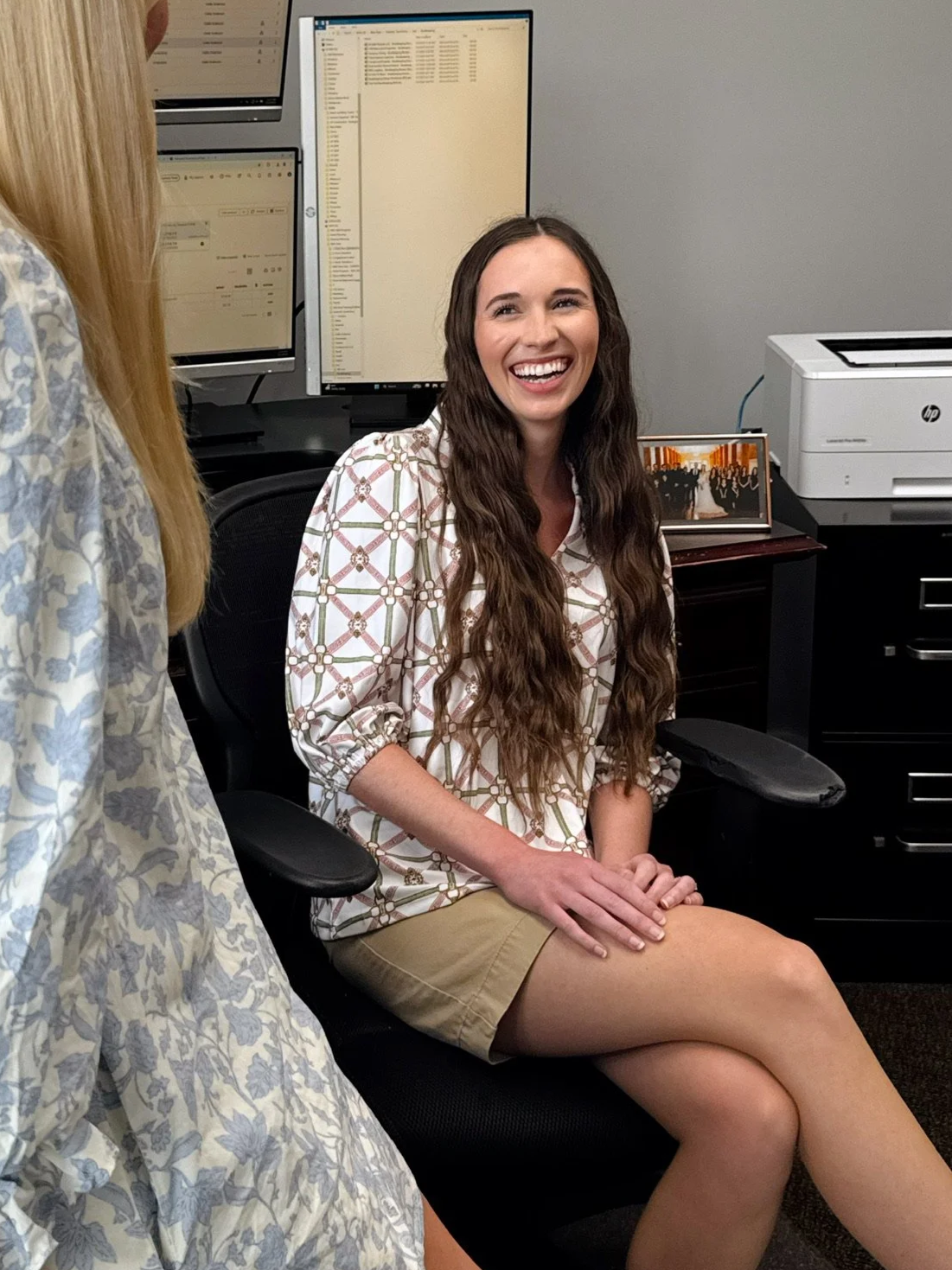 A woman with long curly brown hair smiling and sitting in an office chair in front of dual computer monitors and a printer.