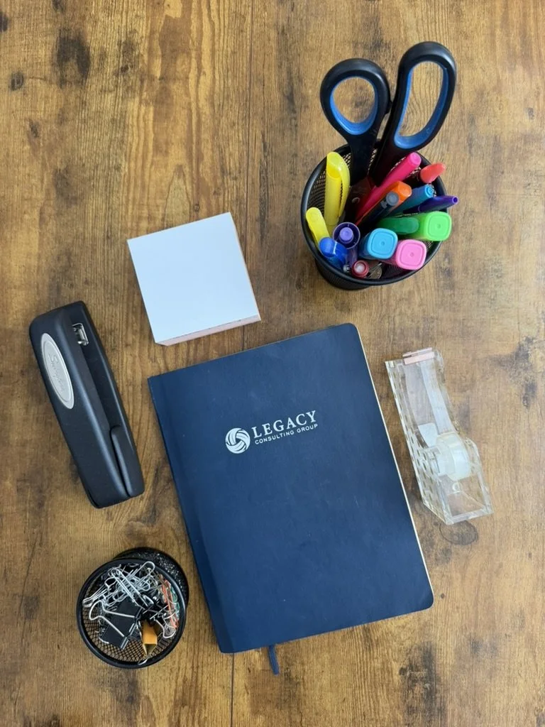 Desk with a blue planner, a container of colorful markers, scissors, a tape dispenser, a sticky note pad, a stapler, and a small container of paper clips.