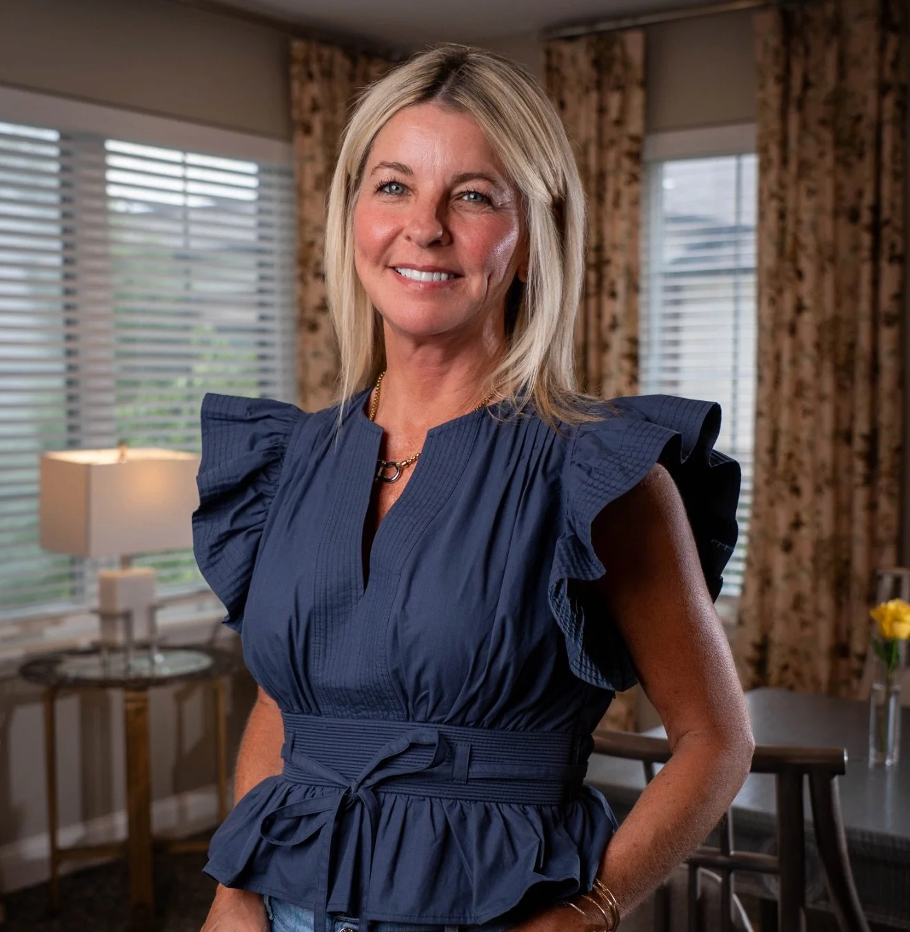 A woman with shoulder-length blonde hair smiling, wearing a navy blue dress with ruffled sleeves, standing in a well-lit room with large windows and patterned curtains.