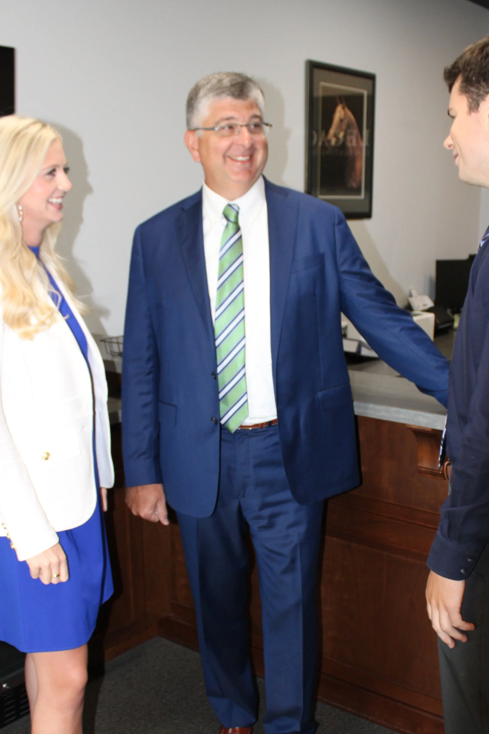 Three people in business attire shaking hands in an office setting, with a framed picture on the wall in the background.