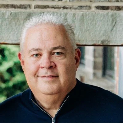 Portrait of a middle-aged man with short white hair, wearing a dark jacket, standing outdoors near a stone wall with blurred greenery in the background.
