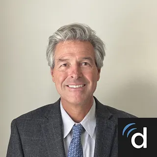 A smiling middle-aged man with gray hair wearing a dark gray suit, white shirt, and blue patterned tie, standing against a plain light-colored background.