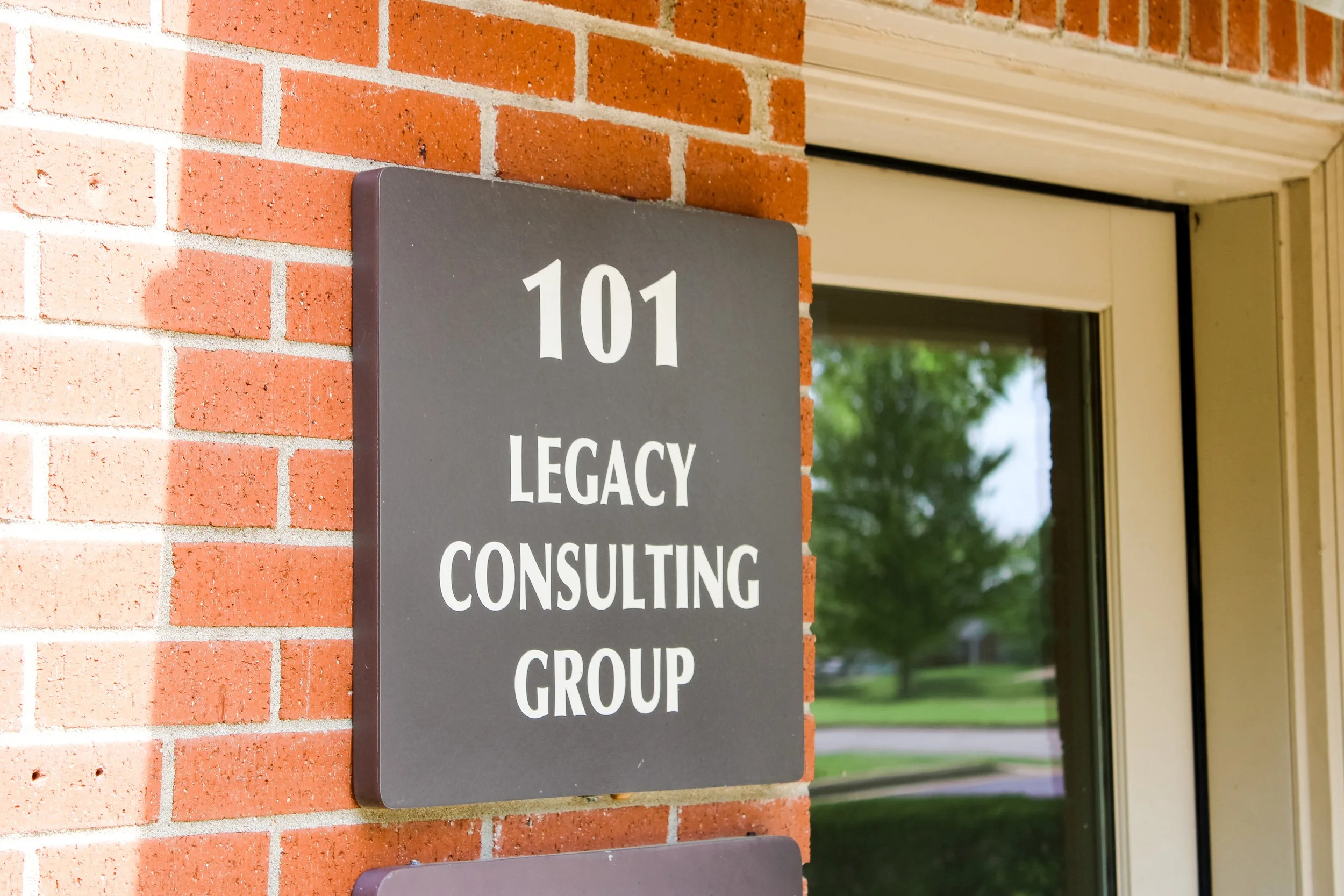 Exterior view of a red brick building with a door and window, showing a gray sign with white text that reads '101 Legacy Consulting Group'.