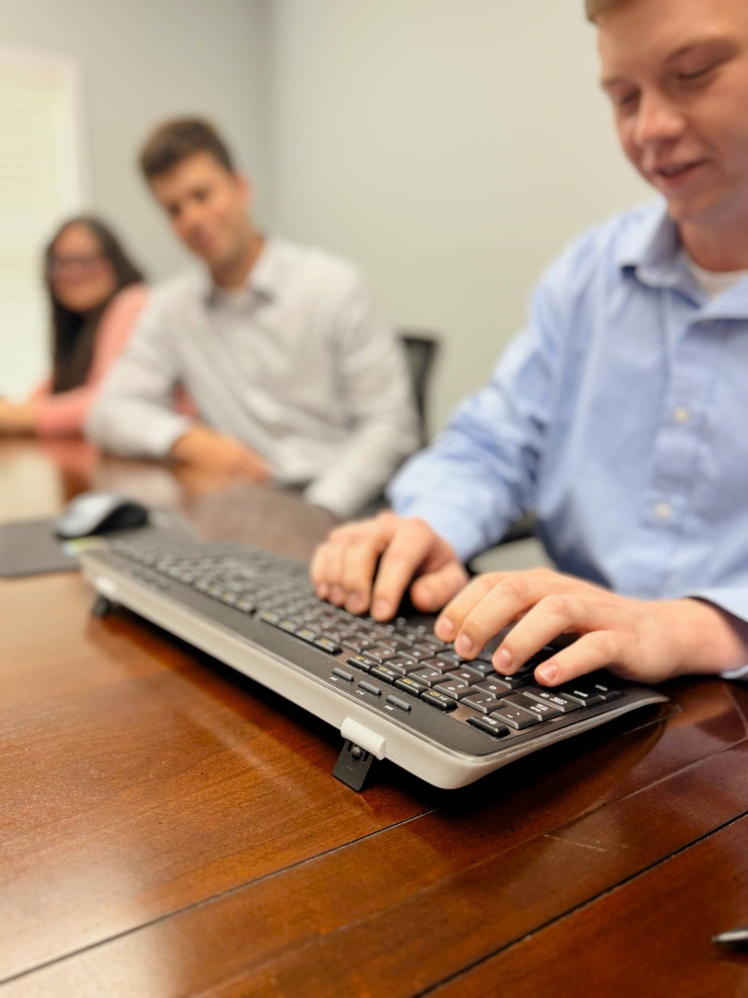 A close-up of a person typing on a computer keyboard at a wooden desk with three other individuals in the background, out of focus.