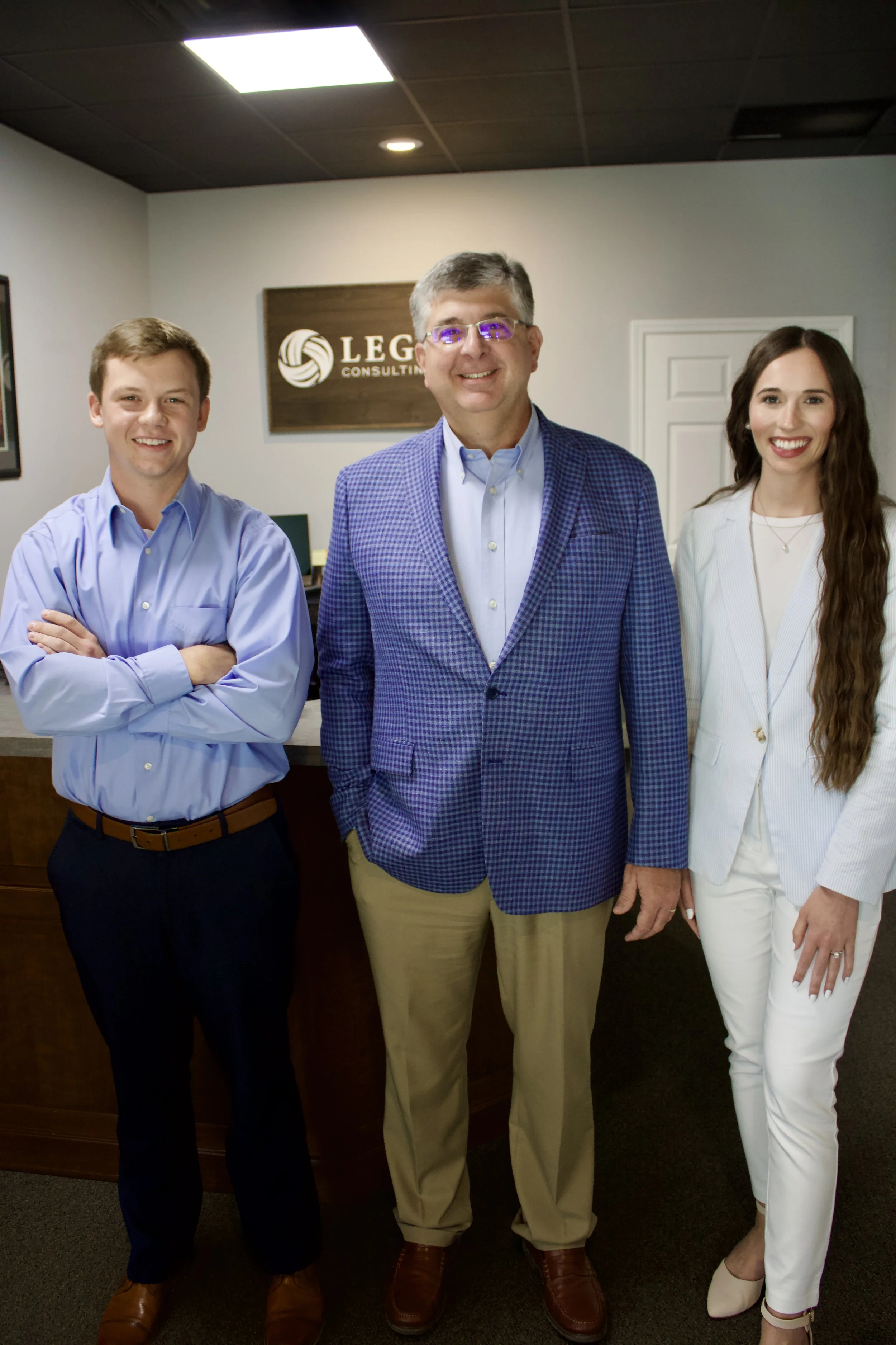 Three professionally dressed people standing together indoors, smiling at the camera. Behind them is a wall sign that reads 'LEGAL CONSULTING.'