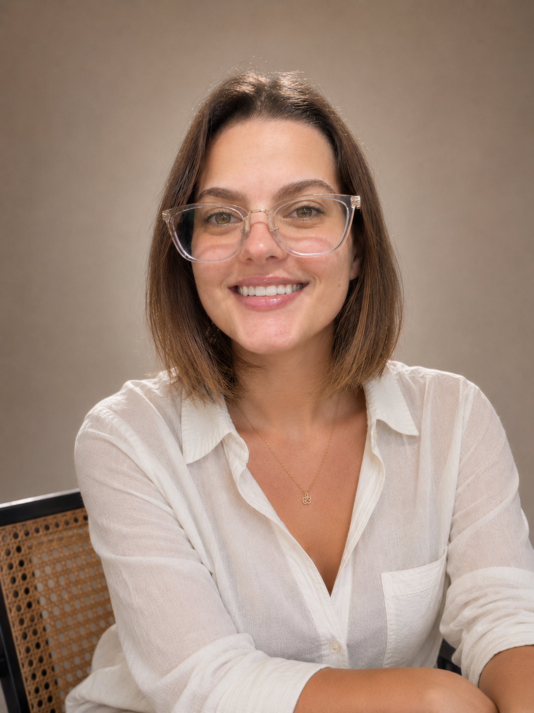 A woman with shoulder-length brown hair wearing glasses and a white button-up shirt, smiling at the camera against a plain background.
