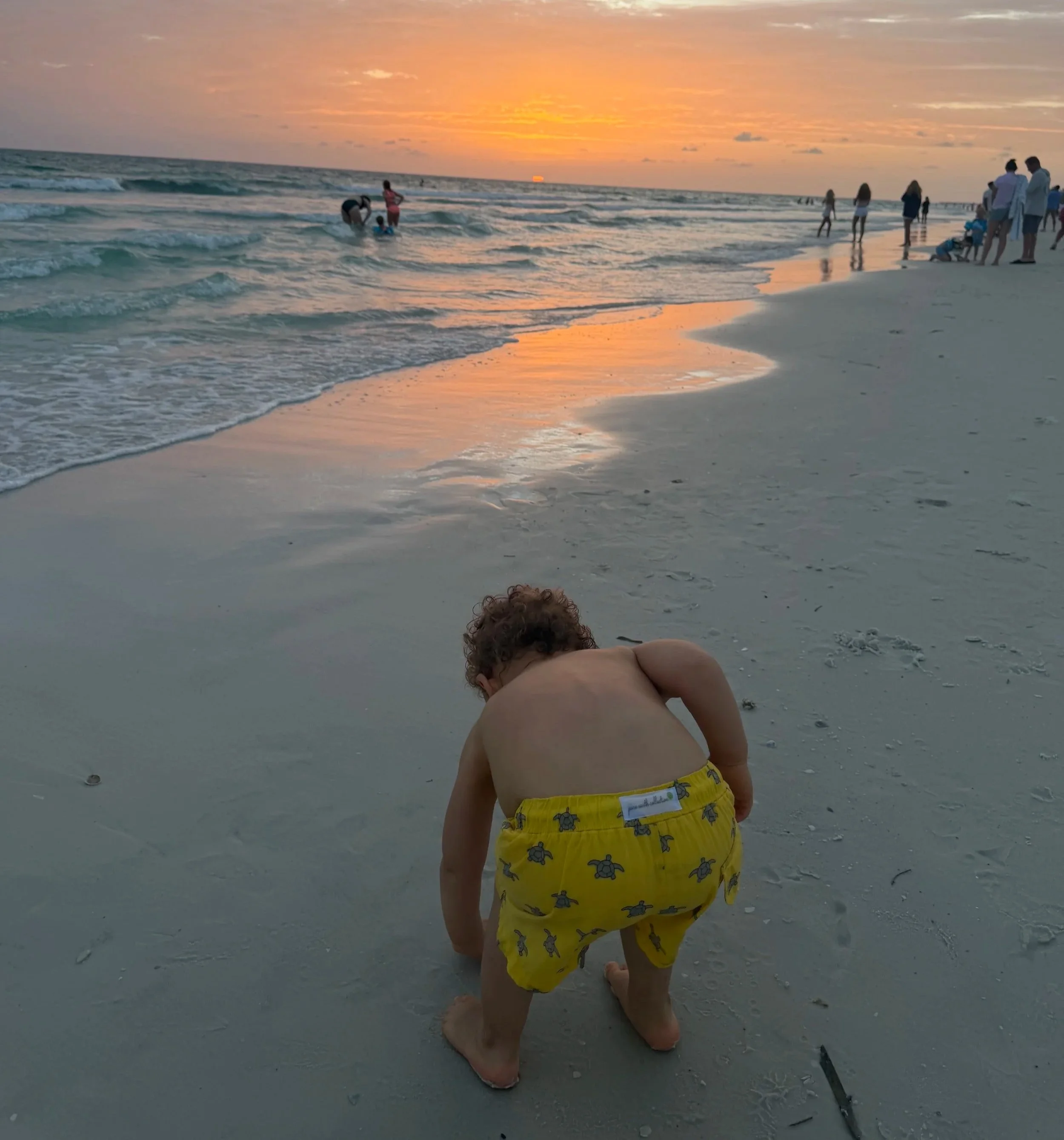 A young child in yellow shorts with turtle patterns is crouching on wet sand at the beach during sunset. The child appears to be playing or digging in the sand, with their back to the camera. The ocean waves are gently coming ashore, and there are people in the distance enjoying the sunset, some standing and some playing in the water.