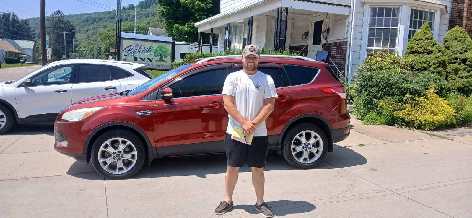 A man standing on a sidewalk in front of a parked red SUV, holding some papers, with a white house and a sign for Big Oaks Inn in the background.