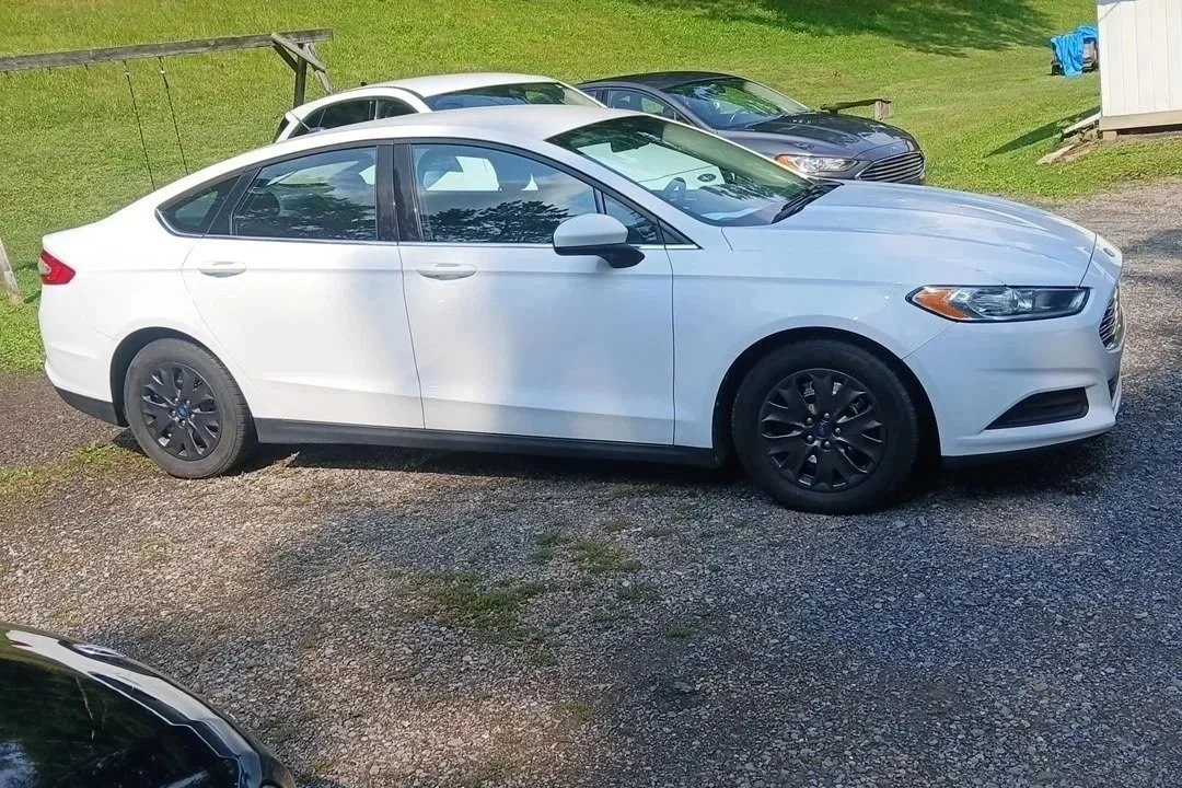 A white sedan car parked on a gravel driveway with other cars in the background, a grassy area, and a small white building.