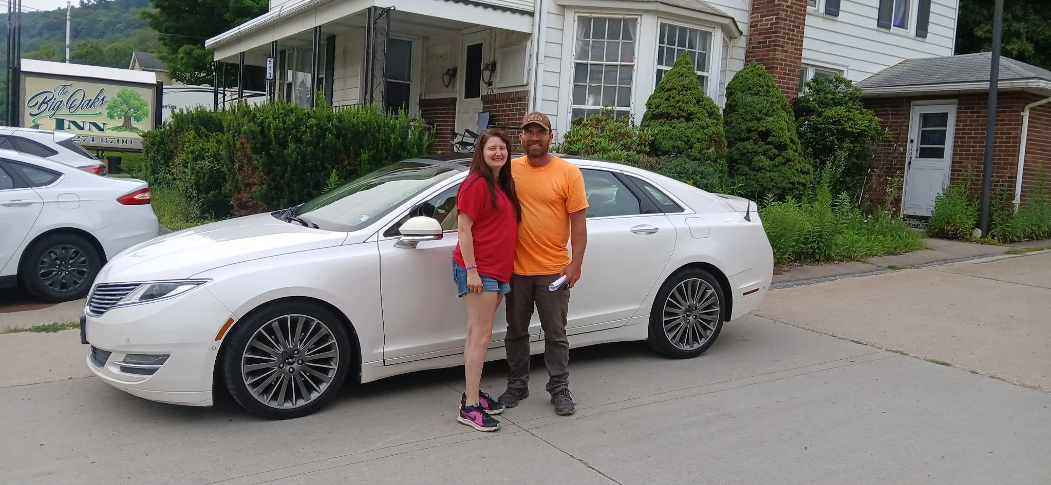 A man and woman standing together outdoors near a white car, smiling, with residential houses and greenery in the background.