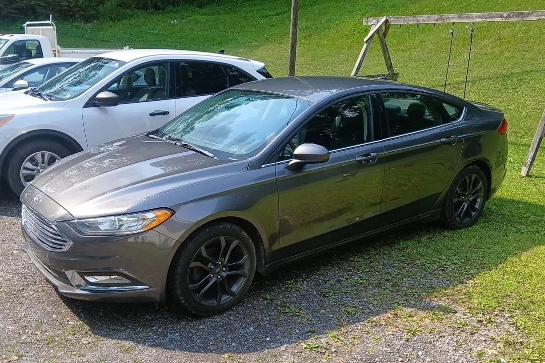 A gray sedan parked next to a white SUV on a gravel surface with a grassy hill in the background.