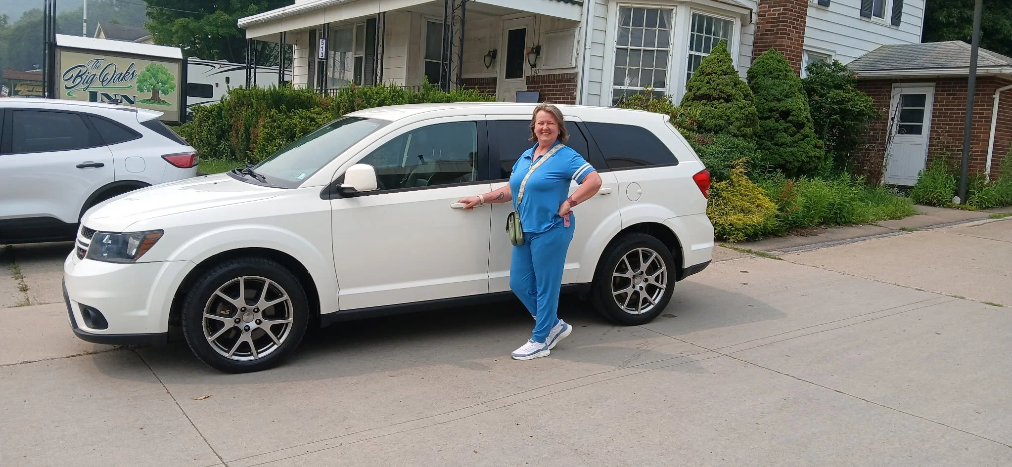 A woman with a big smile wearing blue scrubs, standing next to a white SUV on a residential street.