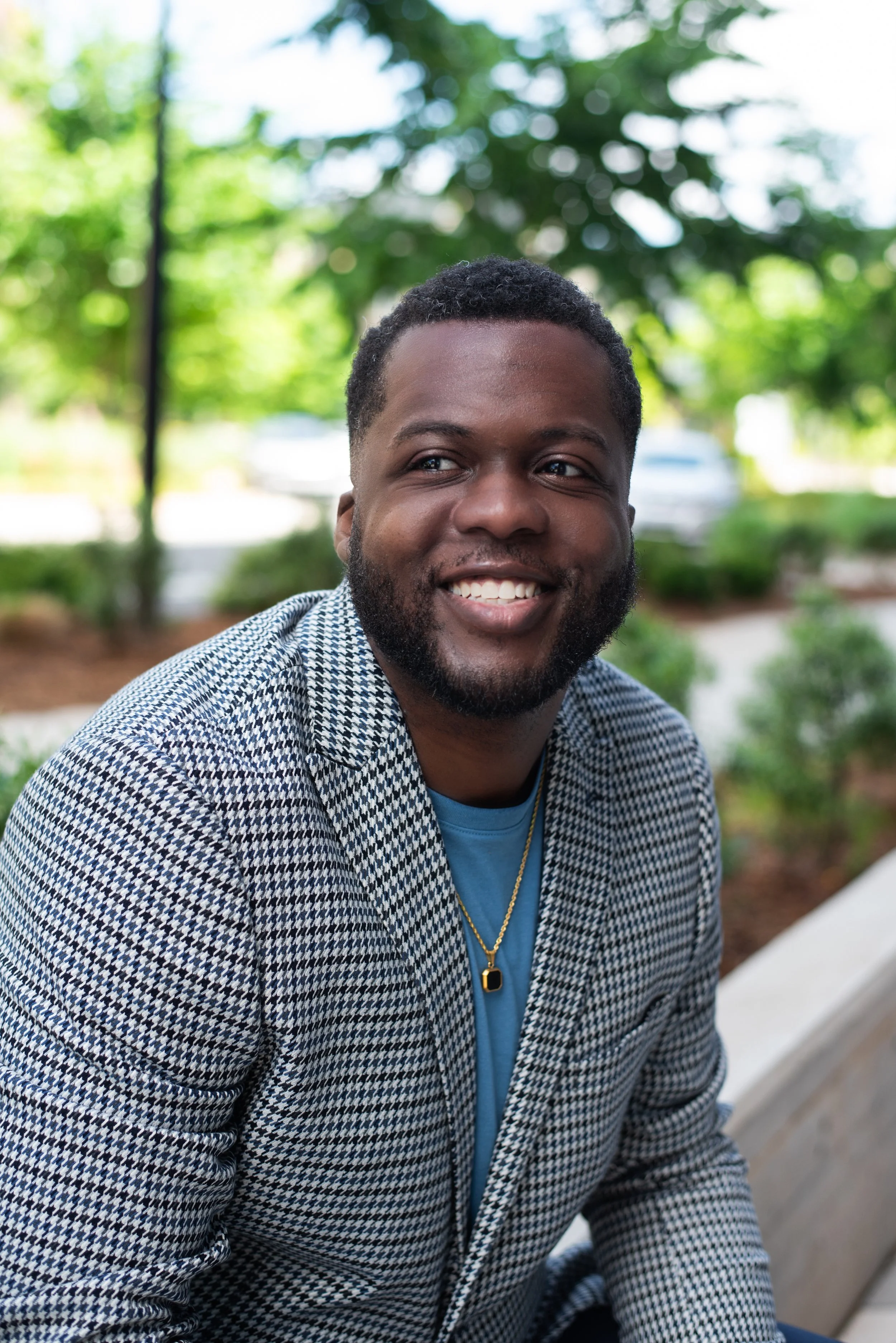 A smiling man with a beard and short curly hair, wearing a patterned jacket, blue shirt, and gold chain, sitting outdoors with green trees in the background.