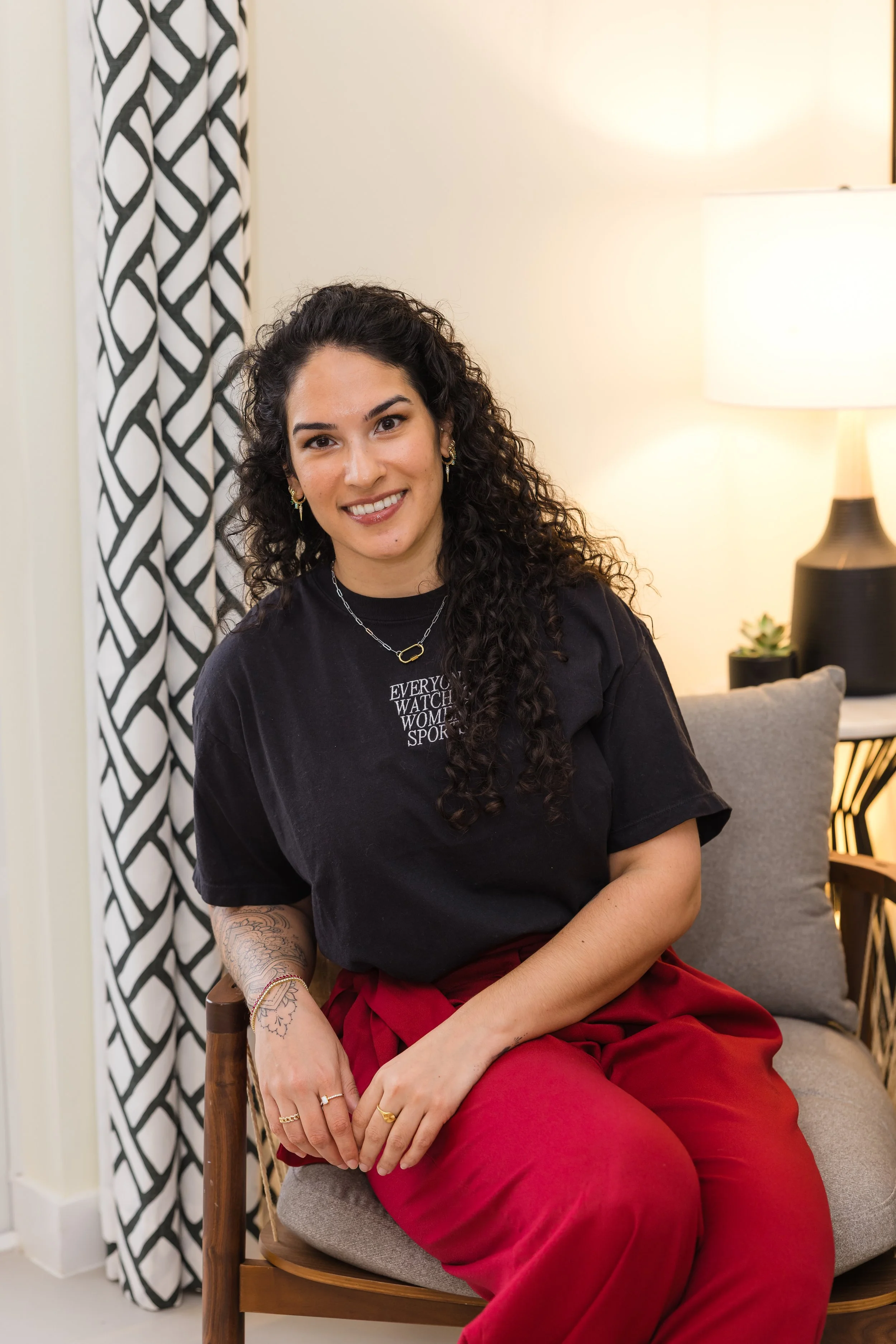 A young woman with curly dark hair, wearing a black T-shirt and red pants, sitting on a chair, smiling in a cozy room with a patterned black and white curtain, a lamp, and a small potted plant in the background.