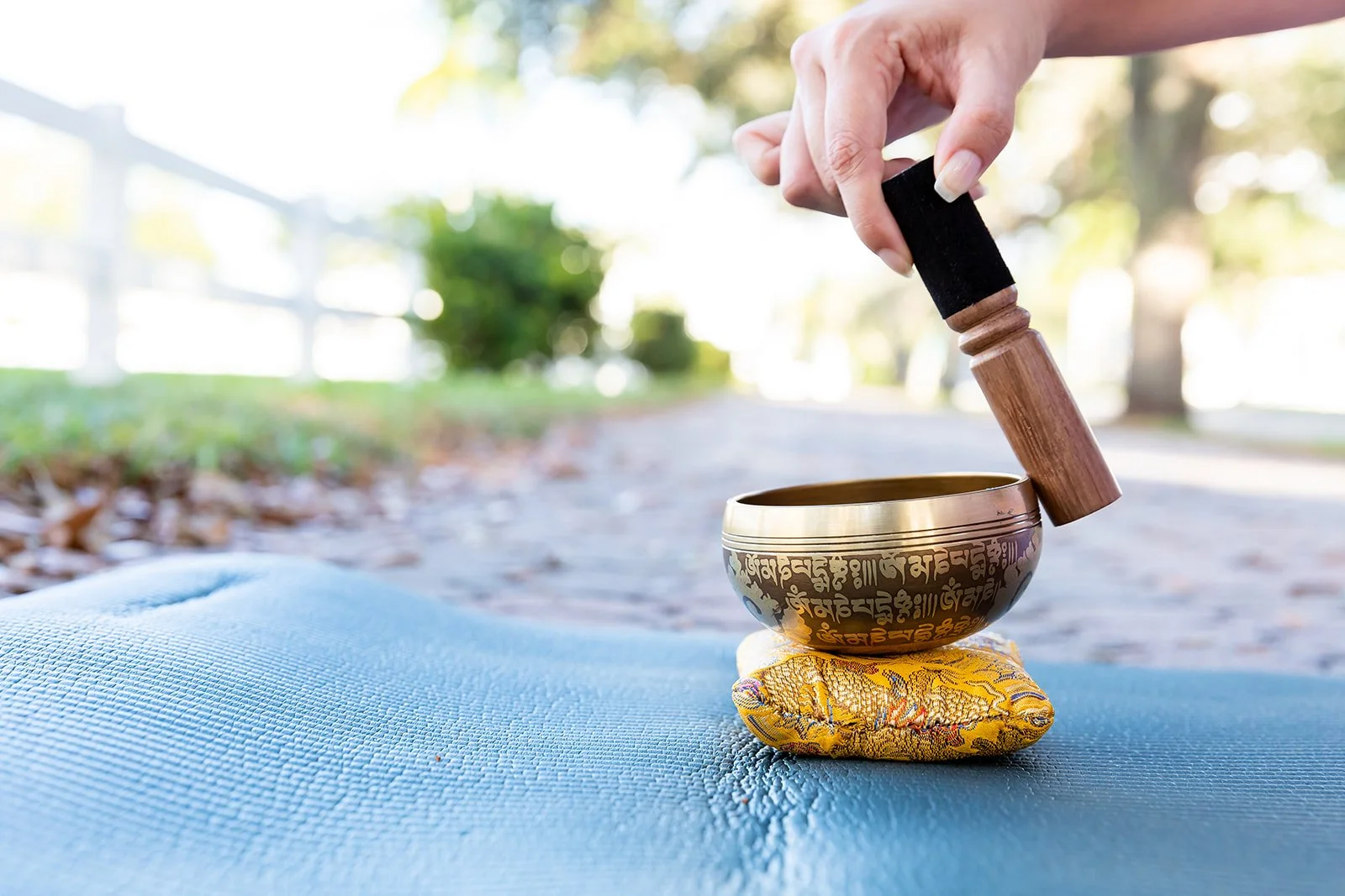 A Tibetan singing bowl with a wooden mallet resting on a yellow cloth, placed on a blue yoga mat outdoors. The background shows trees and a bright sky.