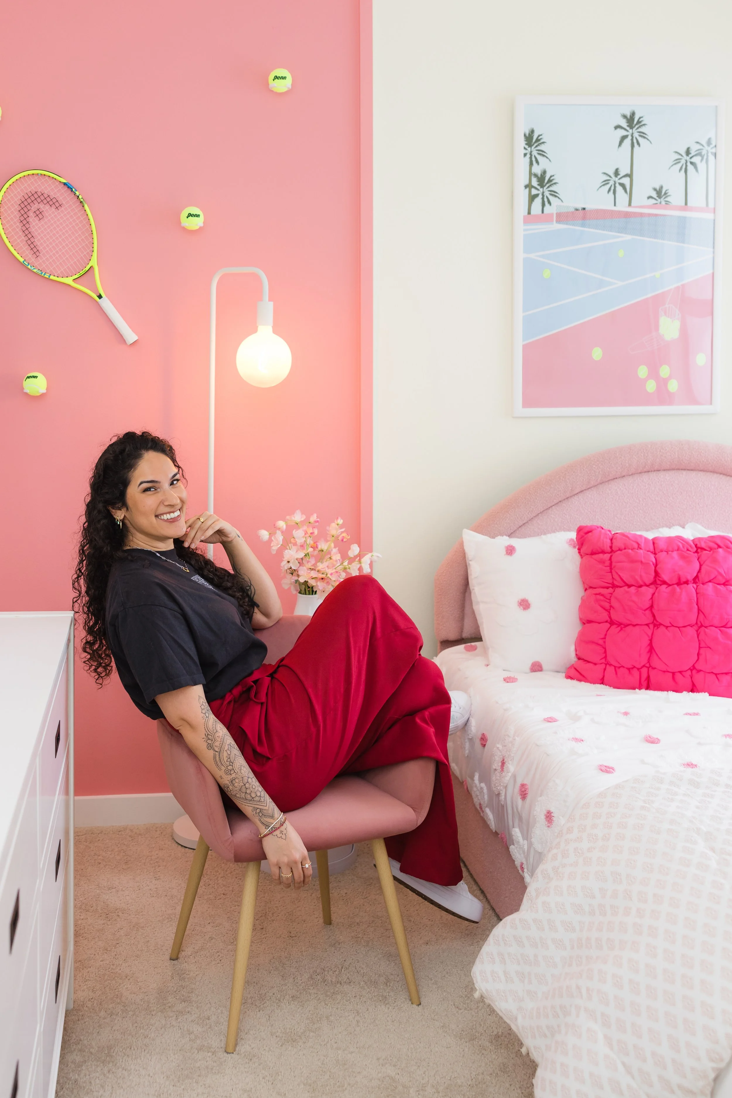 Woman with long curly hair sitting on a pink chair in a pink and white bedroom, smiling at the camera. She is wearing a dark top and red pants, showing tattoos on her left arm. The room has pink and white walls, tennis-themed decor including yellow tennis balls and a tennis racket, framed picture of a tennis court with palm trees, and a bed with a pink, heart-shaped pillow.