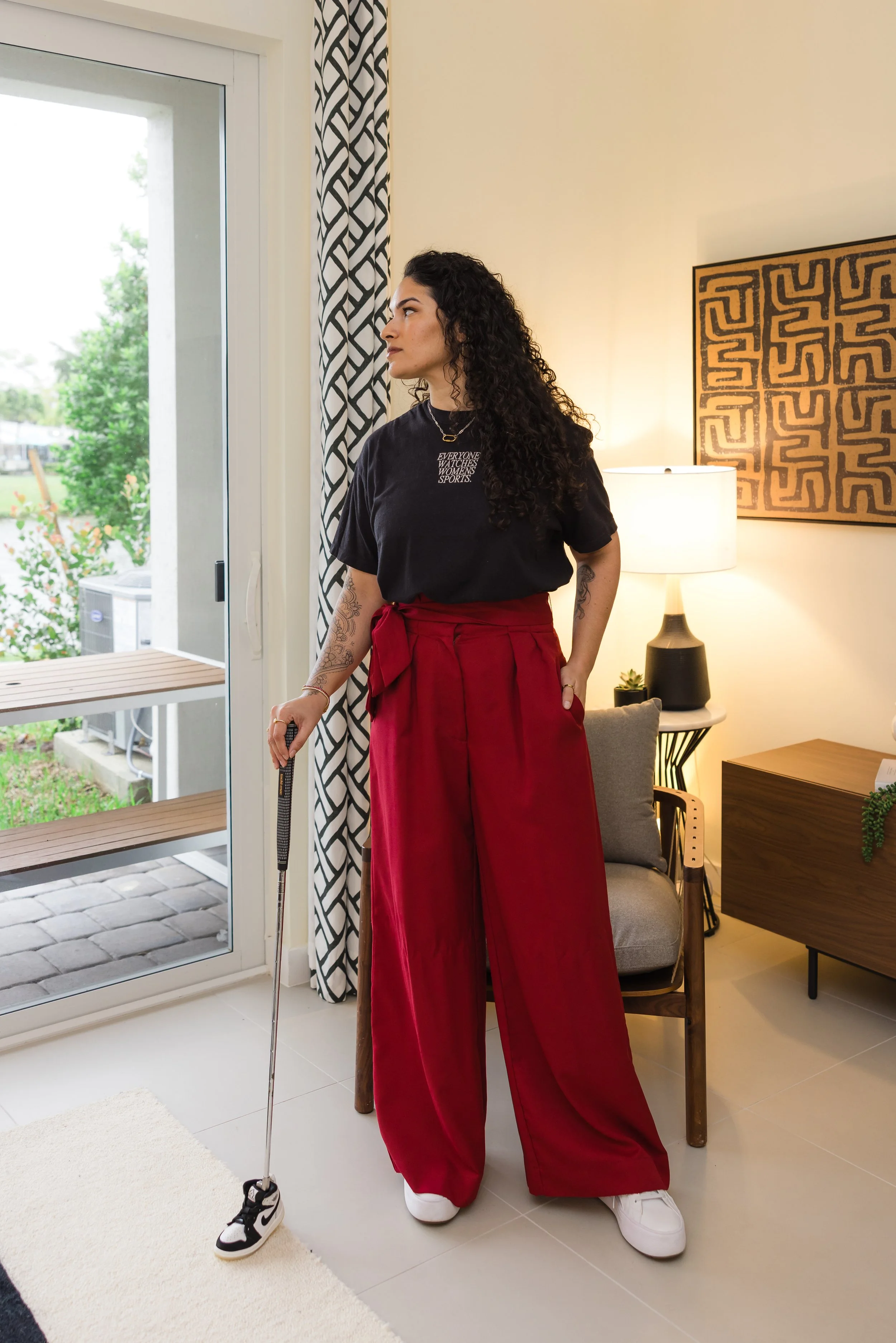 A woman with curly dark hair standing inside of a room, looking to her left, holding a cane, with one foot on a sneaker, and wearing oversized red pants, a black t-shirt, and white sneakers. There is a window behind her with a patterned black-and-white curtain, a lamp, art on the wall, and a small side table with a potted plant.
