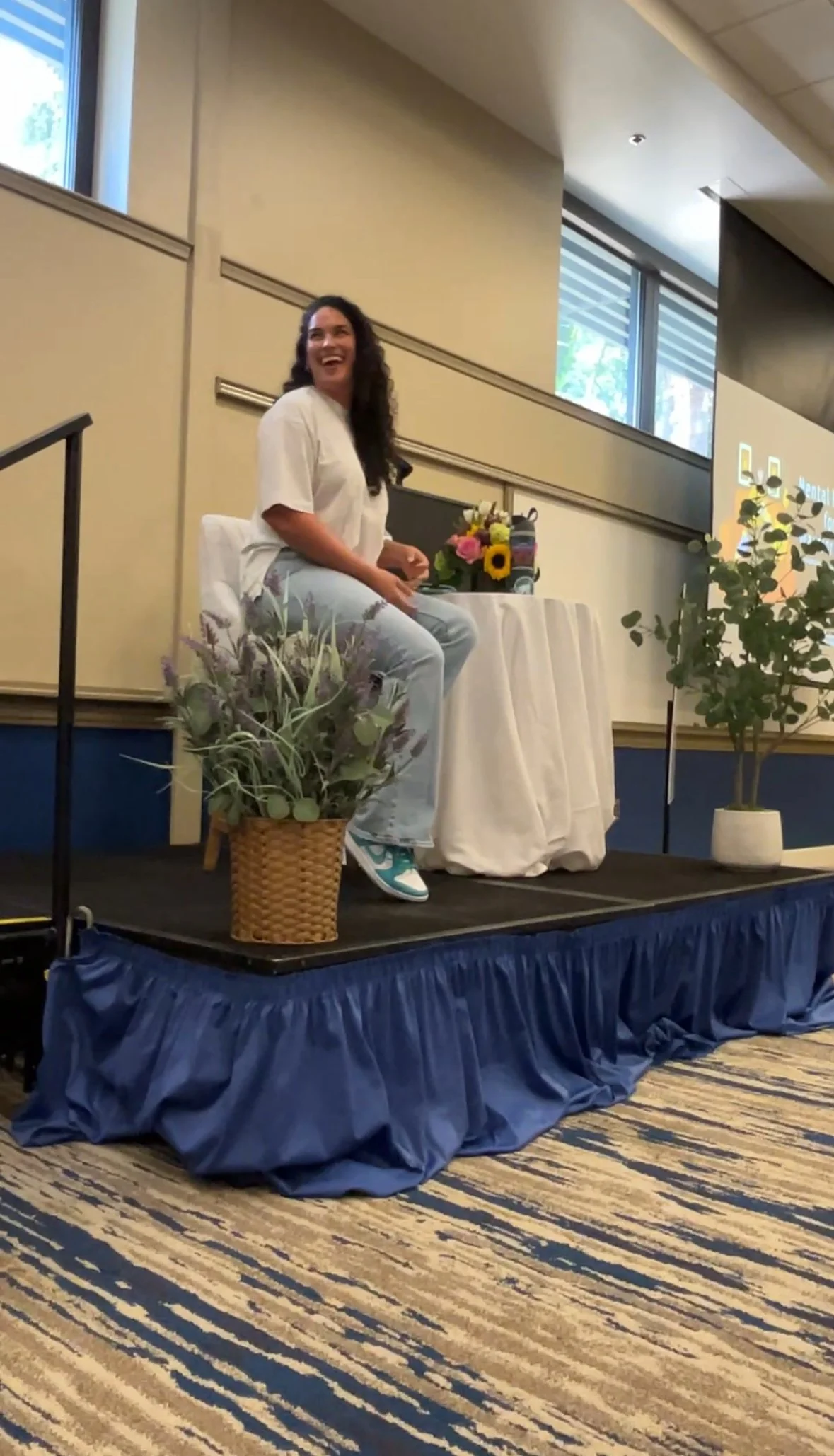 A woman with dark curly hair wearing a white top and light blue jeans, sitting on a stage. There are flowers in a basket and on a table behind her, with a background including a window and a screen.
