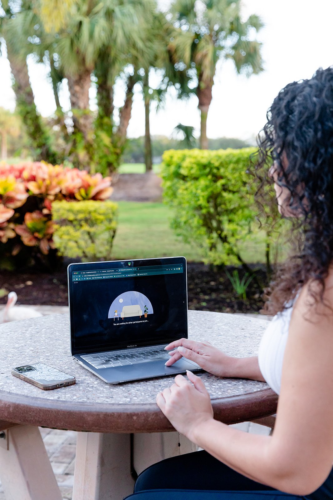 A woman with curly hair sitting at a round outdoor table using a laptop. There is a smartphone on the table, and the background shows a garden with palm trees, bushes, and a grassy area.