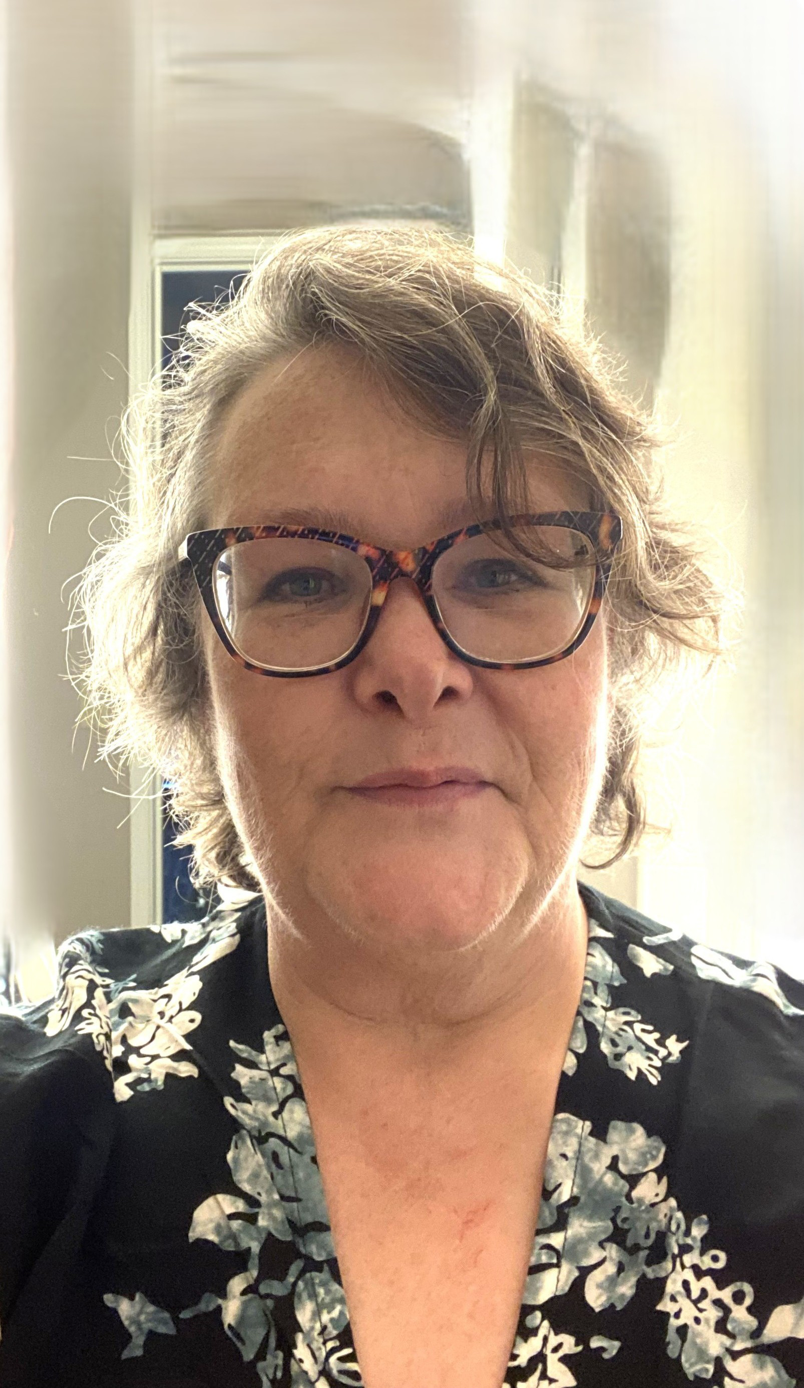 Christine Schut's headshot, a middle-aged woman with short, wavy hair and large, tortoiseshell glasses. She is wearing a black dress with white floral patterns. The background shows a door and light-colored walls, indicating an indoor setting.