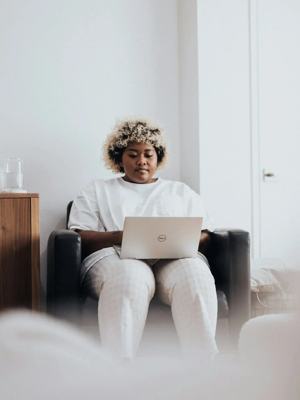 A woman sitting in a black armchair working on a silver Dell laptop in a room with white walls and a small wooden side table with a glass of water on it.