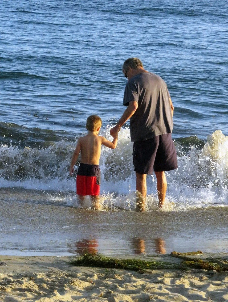 Older man and young boy holding hands at the beach, standing in the shallow water, with waves and sand visible.