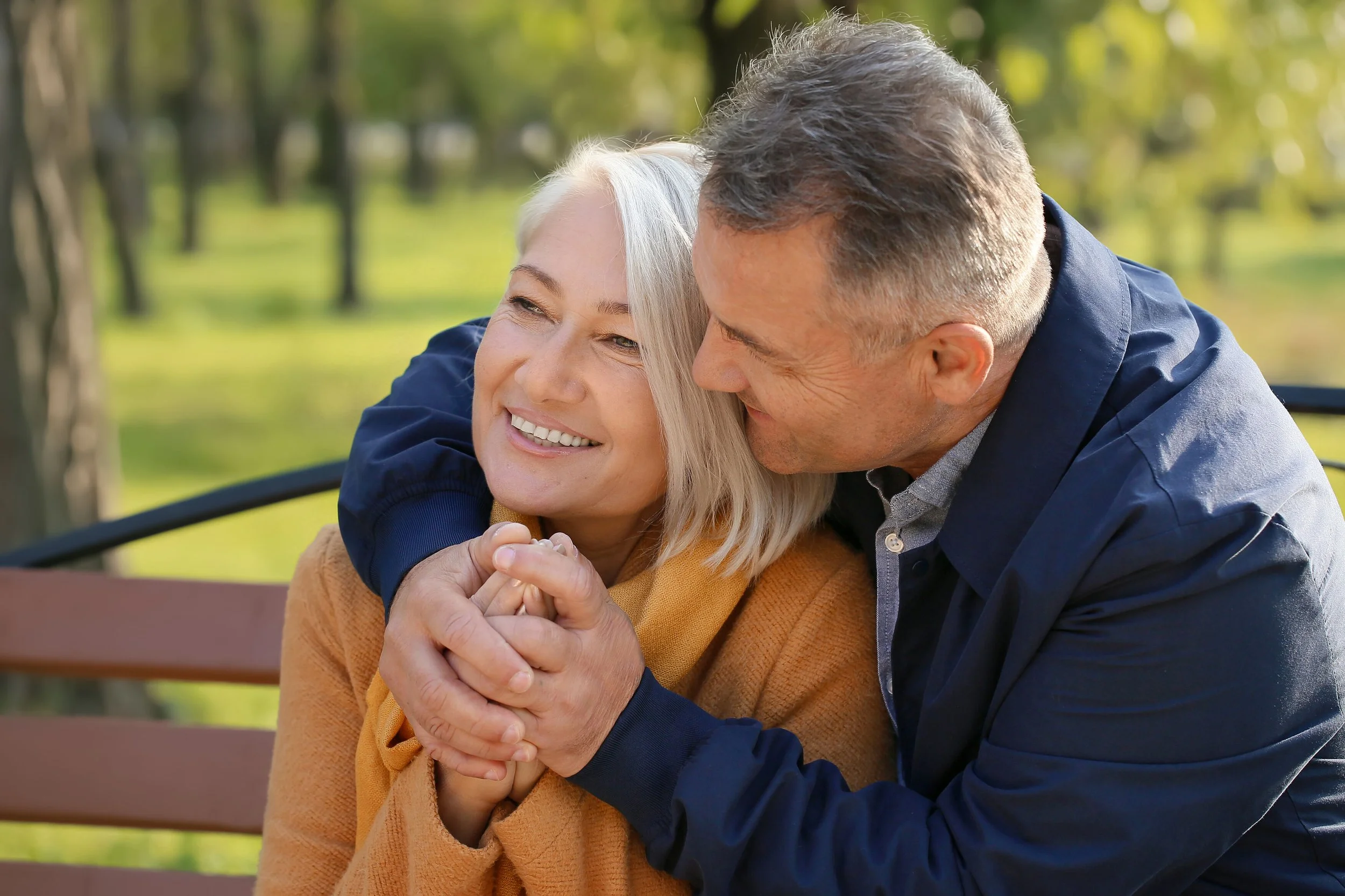 An elderly couple sitting on a park bench, holding hands, sharing a joyful moment, surrounded by green trees and sunlight.