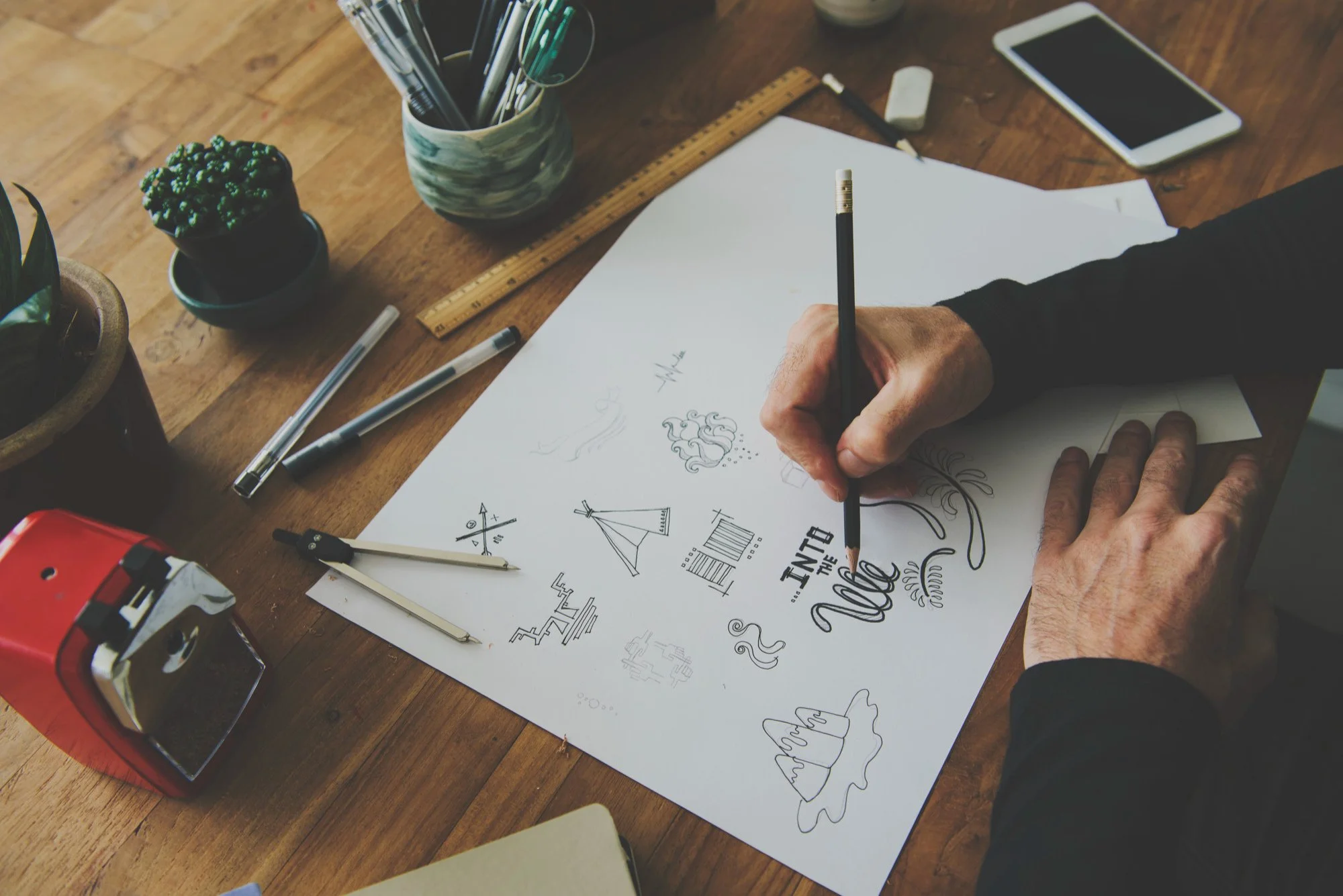 Person sketching on a large sheet of paper with various designs, illustrations, and handwritten text, surrounded by markers, pens, ruler, phone, and plants on a wooden desk.