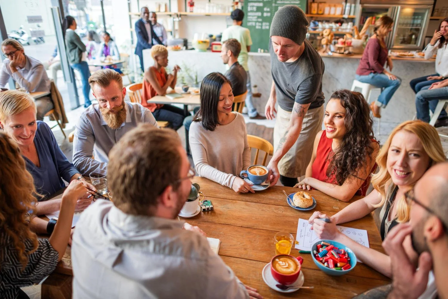 A group of friends gathered around a wooden table in a cozy cafe, enjoying coffee and snacks, with others socializing inside and outside in the background.