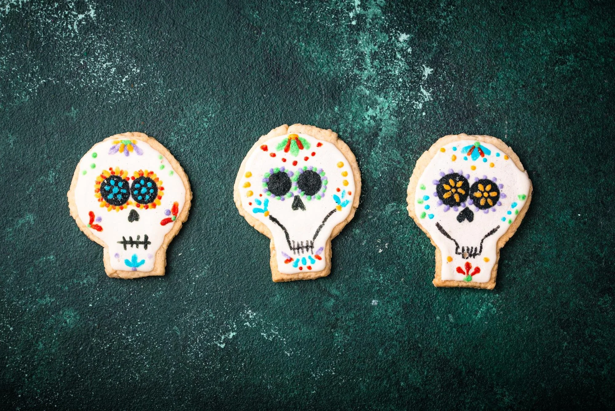 Three sugar cookies decorated with colorful sugar icing in the shape of sugar skulls, placed on a textured dark green surface.
